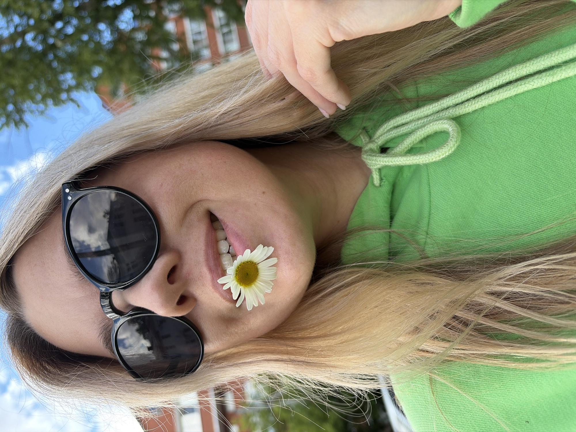 A cheerful blonde woman is depicted in a close-up, angled outdoor portrait in Chaikovsky, Russia. Her long, straight hair cascades around her face and shoulders. She is wearing a bright lime green hooded sweatshirt, with the drawstring visible on her right side. Her eyes are covered by stylish dark, round sunglasses, which reflect a bright sky with white clouds and the surrounding environment. A small, white daisy flower with a prominent yellow center is playfully held between her lips, resting on her bottom lip, revealing a hint of a smile and her white teeth. Her right hand, with light pink fingernails, is visible near her head, gently resting on her hair. The background is softly blurred, indicating an outdoor setting with green foliage or trees against a blue sky, and some distant red-brick buildings with white window frames. The image conveys a relaxed and joyful mood under clear daytime conditions. No text is visible in the media file.