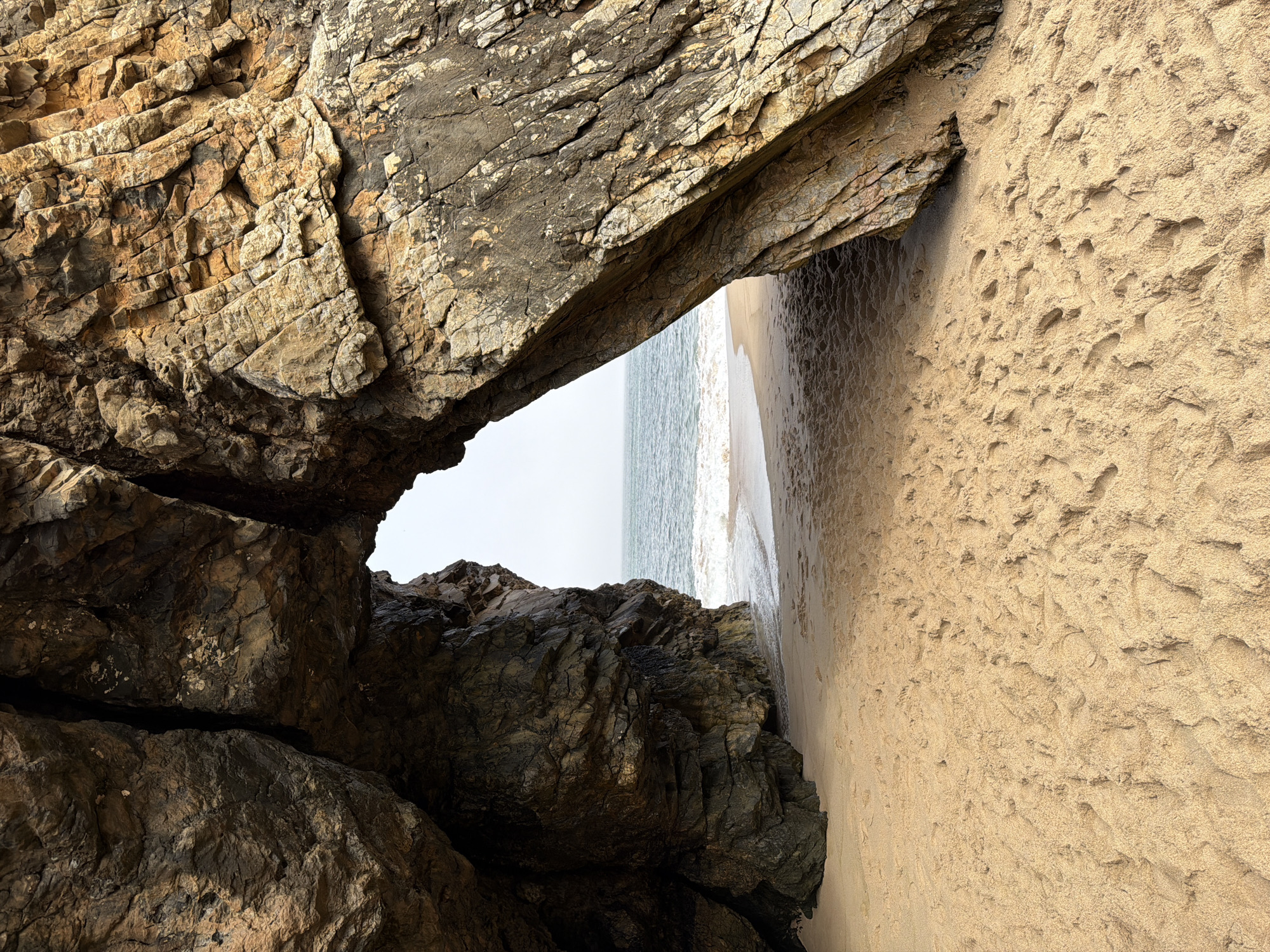 This is a photograph taken from within a sea cave at Praia da Adraga, Colares, Portugal. The cave opens to reveal a sandy beach with the Atlantic Ocean beyond. The rough texture of the cave walls, composed of layered rock formations in shades of brown and grey, are visible on both the left and right sides of the frame. The sand on the beach is light brown and shows numerous footprints. The ocean water is blue-green with small waves breaking on the shore. The sky is overcast. There are no people or man-made objects visible in the scene.