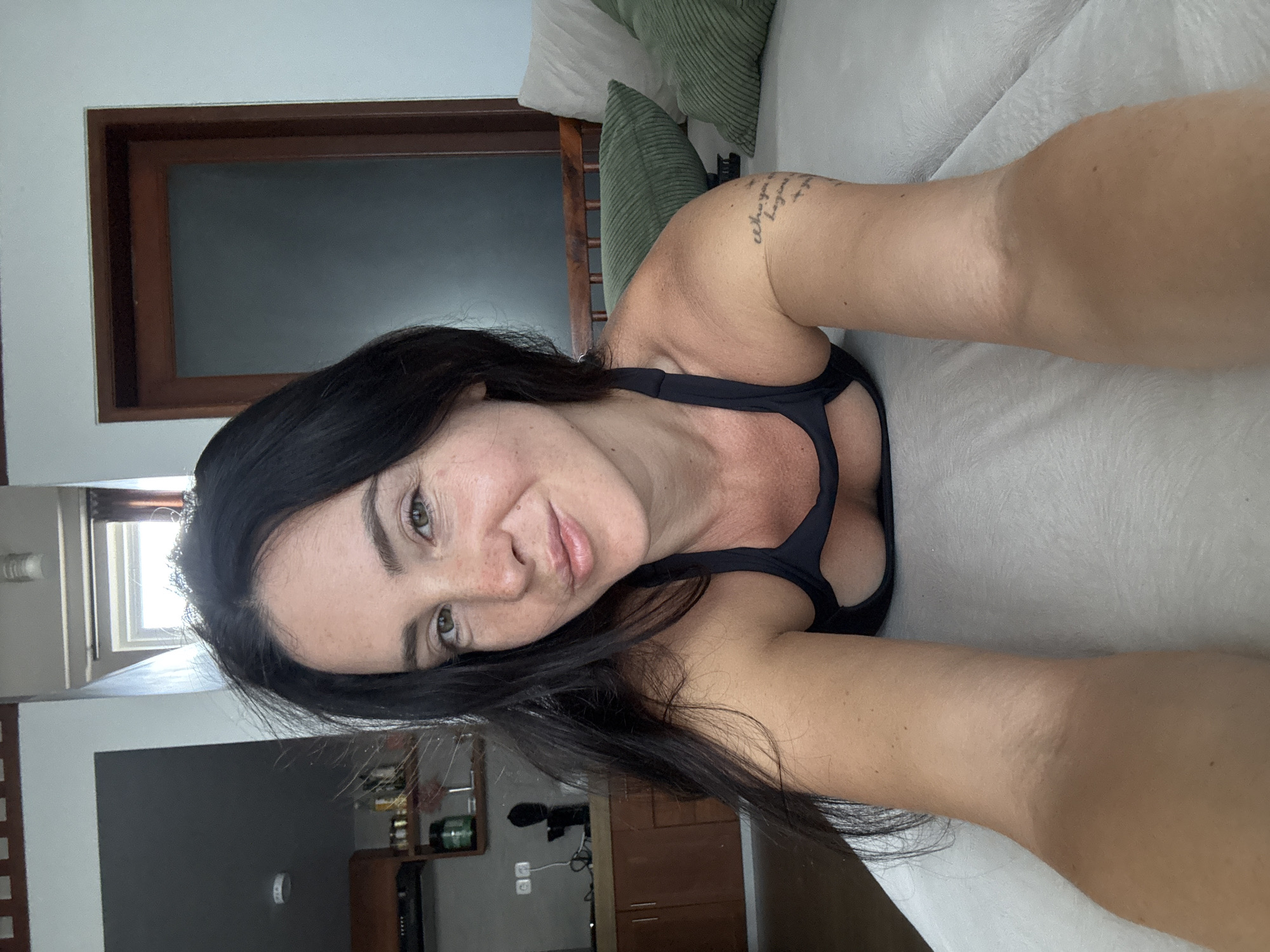 A woman with dark hair and freckles is lying on a white bed, looking at the camera. She is wearing a black sports bra. In the background, there is a kitchen area with wooden cabinets and shelves with bottles and containers. To the right of the woman, there are green and white pillows on the bed and a wooden railing. A wooden door with a frosted glass panel is visible in the background. The location is an indoor setting, possibly a bedroom or living area in North Kuta, Indonesia.