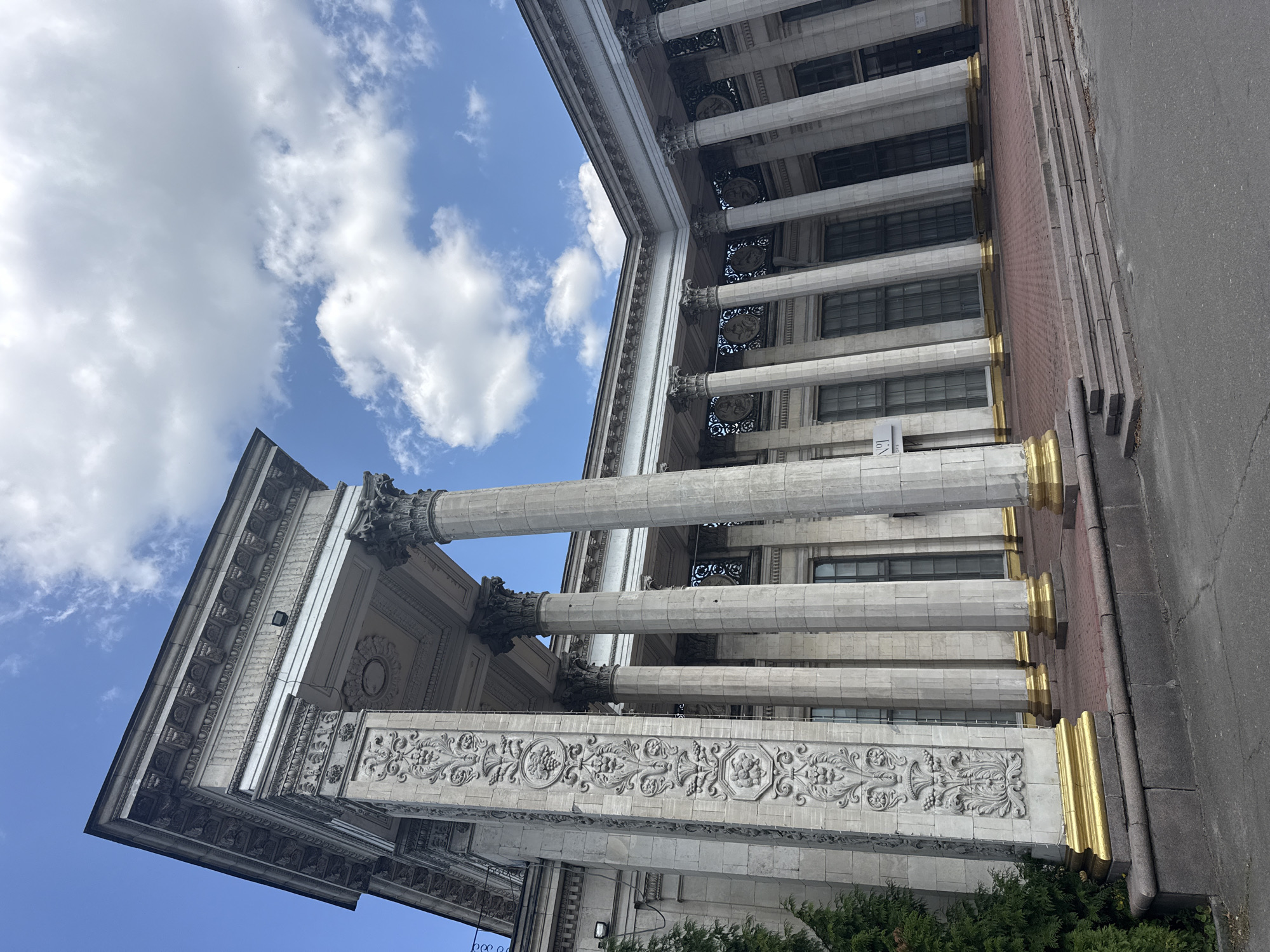 The image captures the imposing facade of the National Academy of Sciences of Ukraine in Kyiv, Ukraine. The building is characterized by its neoclassical architecture, featuring a colonnade of tall, fluted Corinthian columns supporting a classical entablature. The columns are made of light-colored stone, with ornate golden bases. Intricate stone carvings adorn the surface of some of the columns, depicting floral motifs and what appear to be mythological figures.
The main facade is a light gray stone, with large, tall windows that are dark-tinted, reflecting the sky and clouds. The building's roofline is adorned with detailed cornices and decorative elements. In the foreground, a broad, stepped entrance leads up to the building, paved with red brick and granite. A large green evergreen tree is visible to the far left of the image, partially obscuring the building's corner.
The sky above is a bright blue, populated by fluffy white cumulus clouds, suggesting a clear and pleasant day. There are no people or animals visible in the scene. The overall impression is one of grandeur, history, and architectural significance.