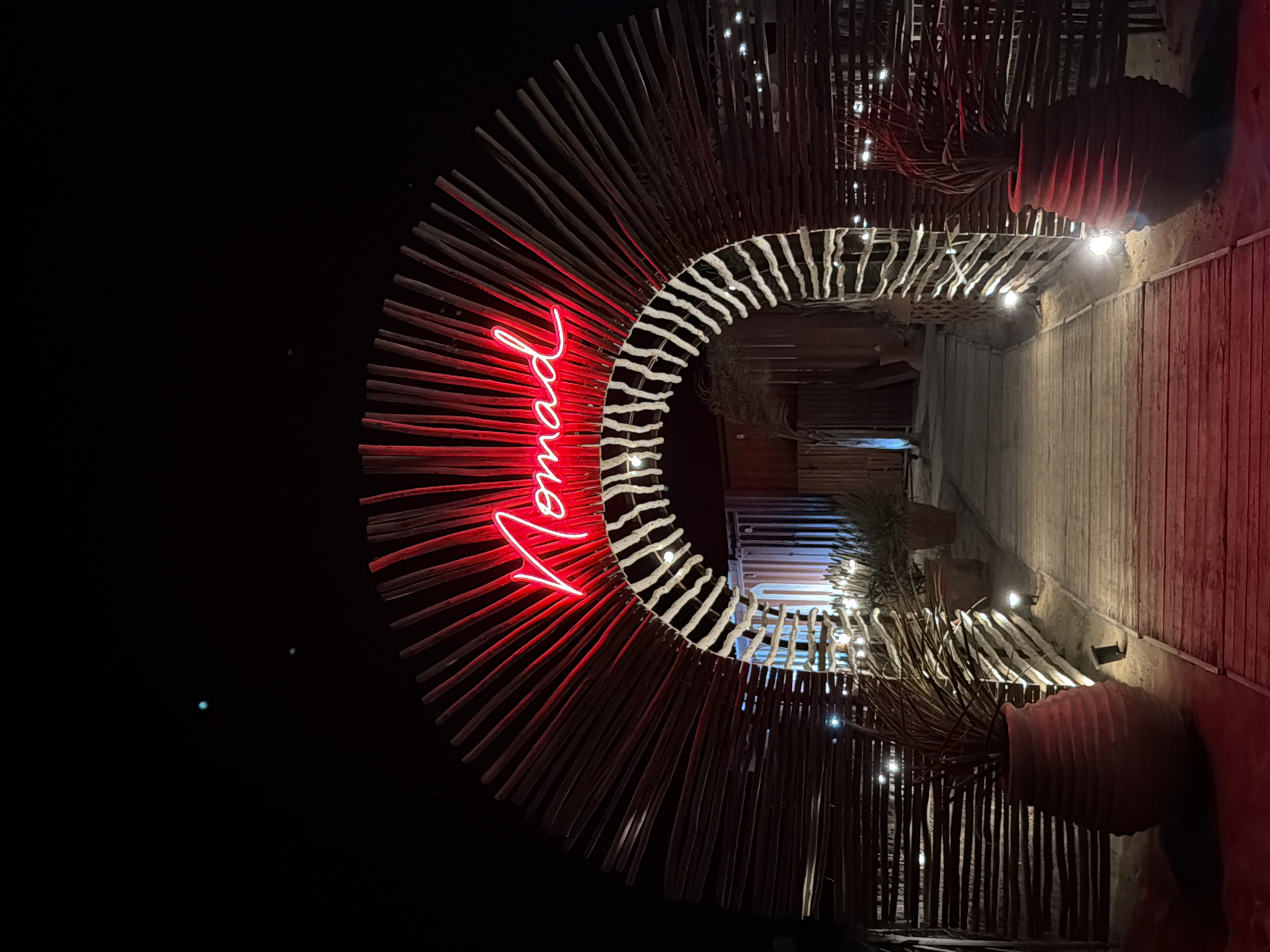 The image depicts an archway constructed from natural wood sticks, illuminated by red neon signage spelling out "Nomad." The archway is set against a dark night sky. A wooden walkway leads through the archway, flanked by large, textured terracotta pots containing sparse vegetation. Small, upward-facing spotlights are embedded along the walkway and the base of the archway, casting a warm glow. The environment appears to be a desert or sandy area, with the ground around the walkway being sandy. In the sky, two small, bright celestial bodies are visible. The overall scene suggests an entrance to an establishment or event, possibly a restaurant or bar, in a bohemian or rustic setting. The location is identified as the Nomad beach bar in Torreilles, France.