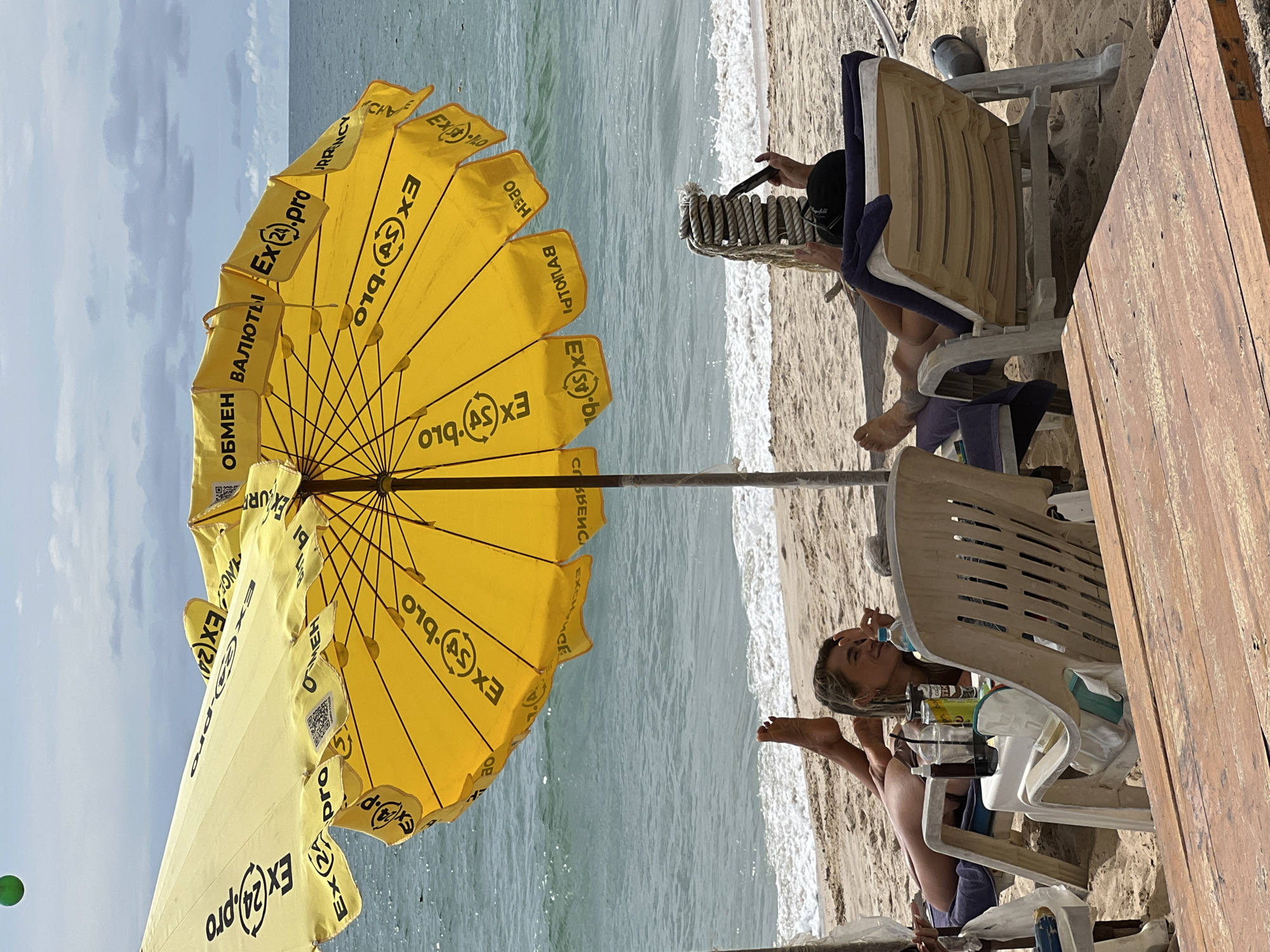 Two people are lounging on beach chairs under a yellow umbrella on a sandy beach. The umbrella is branded with "EX24.pro" and other text. The ocean is visible in the background with waves breaking on the shore. The person on the left is a woman lying on her side, looking towards the camera and smiling. The person on the right is mostly obscured, seen from behind, with their legs propped up and holding a phone. There are drinks and a cooler on a small table between them. In the distance, the sea stretches to the horizon under a cloudy sky. A green ball is visible in the upper left corner of the image.