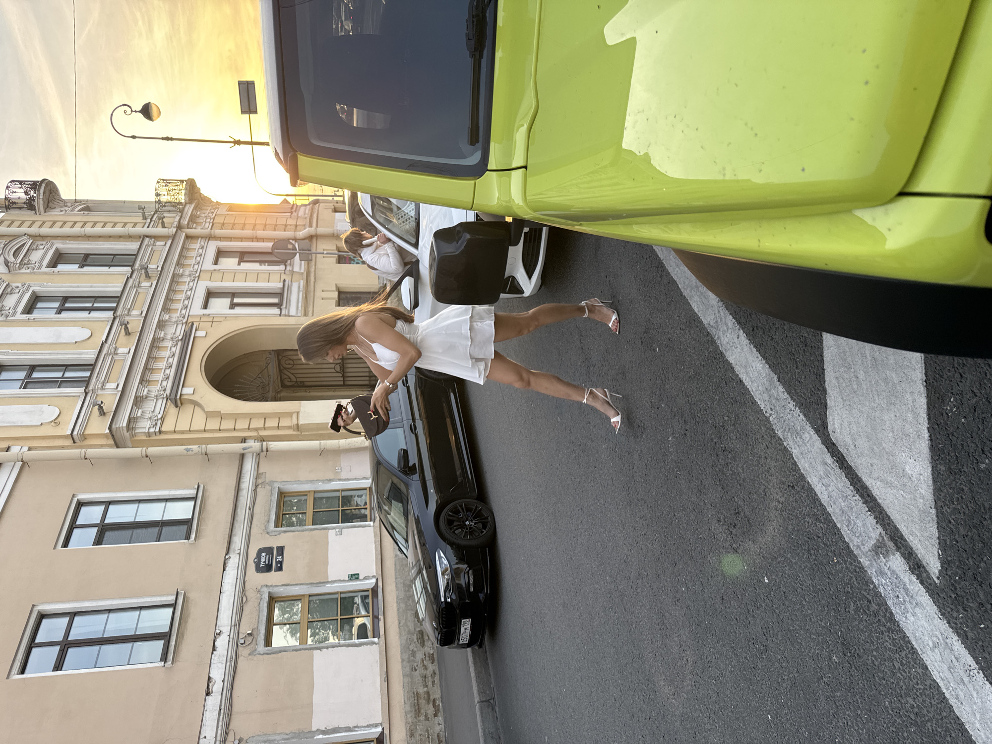 The image depicts a street scene in Saint Petersburg, Russia. In the foreground, a bright yellow SUV is parked, partially obscuring the view. A woman wearing a short white dress and high-heeled sandals is walking away from the camera, holding a dark handbag. She is positioned on the right side of a black BMW sedan. Behind the BMW, another white car is visible, and a person in a white jacket appears to be inside it or standing next to it. The buildings lining the street are of classical architecture, with arched doorways and decorative facades. A street lamp with an ornate design is also visible. The setting sun casts a warm, golden light over the scene, creating long shadows. The street is paved with asphalt, and white lines mark parking spaces. A street sign with Cyrillic lettering, indicating "ТУЧКОВ 24", is attached to one of the buildings, suggesting the location is Tuchkov Lane, number 24. The overall atmosphere is that of a late afternoon or early evening in an urban environment.
