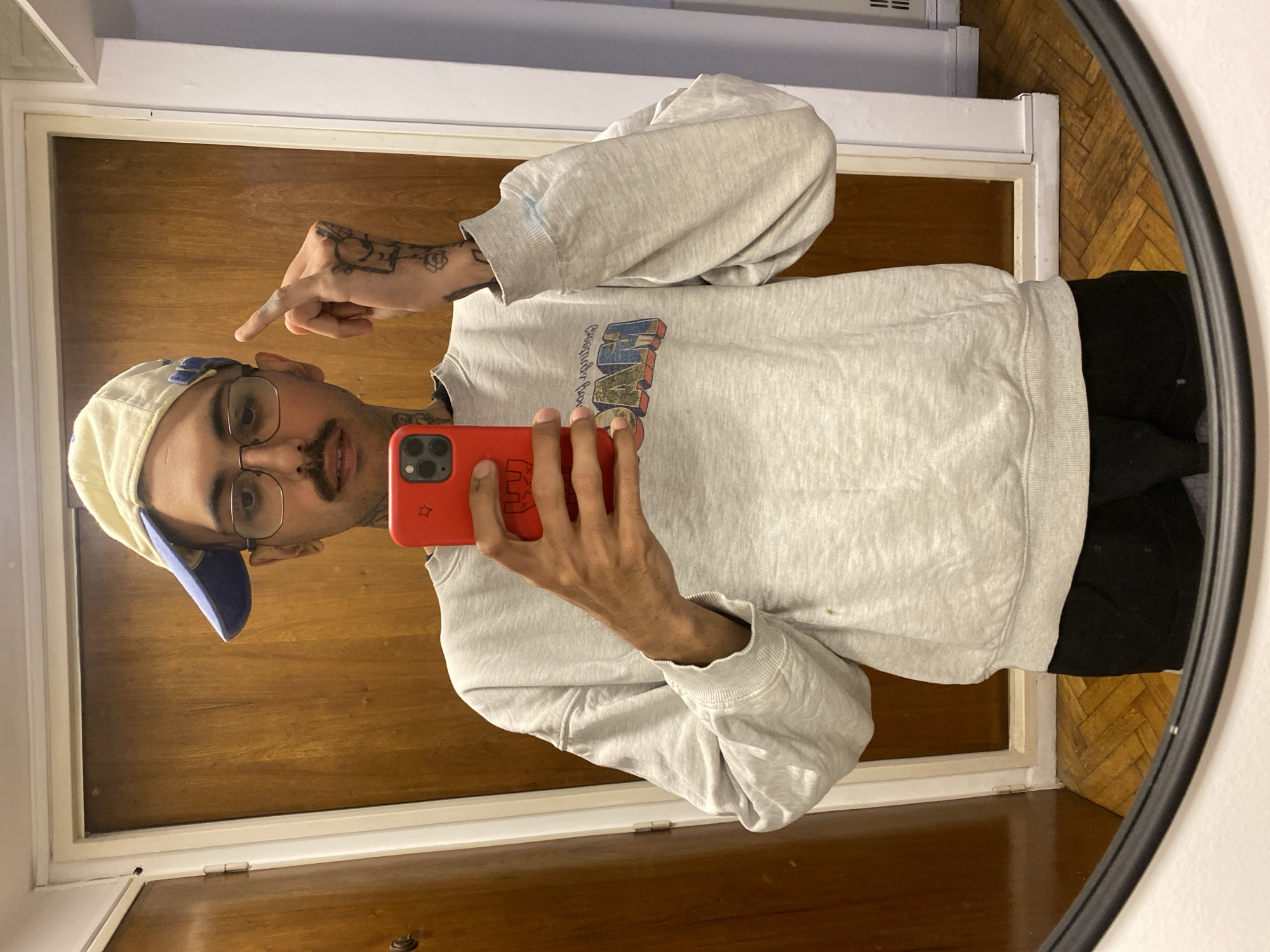 The image captures a vertically oriented mirror selfie of a young man indoors, likely in Buenos Aires, Argentina. The man is reflected from the chest up, wearing a light grey long-sleeved sweatshirt featuring a colorful, three-dimensional graphic on the front that appears to read "CLUB DEPORTIVO" or "ITALIANO." He dons a cream-colored baseball cap with a blue brim and clear-rimmed glasses, complementing his moustache and soul patch. His left hand, visible with various tattoos including a rose and abstract patterns, is raised with the index finger pointing towards his head. He holds a red smartphone, featuring a small white star on its case, in his right hand to capture the selfie.
The reflected background reveals a warm-toned wooden door and a white door frame. A section of a wooden floor with a distinctive herringbone pattern is visible at the bottom right of the reflection. The mirror has a dark, possibly black, frame partially seen on the right. The indoor lighting suggests daytime. The man maintains a neutral expression while looking at his phone.