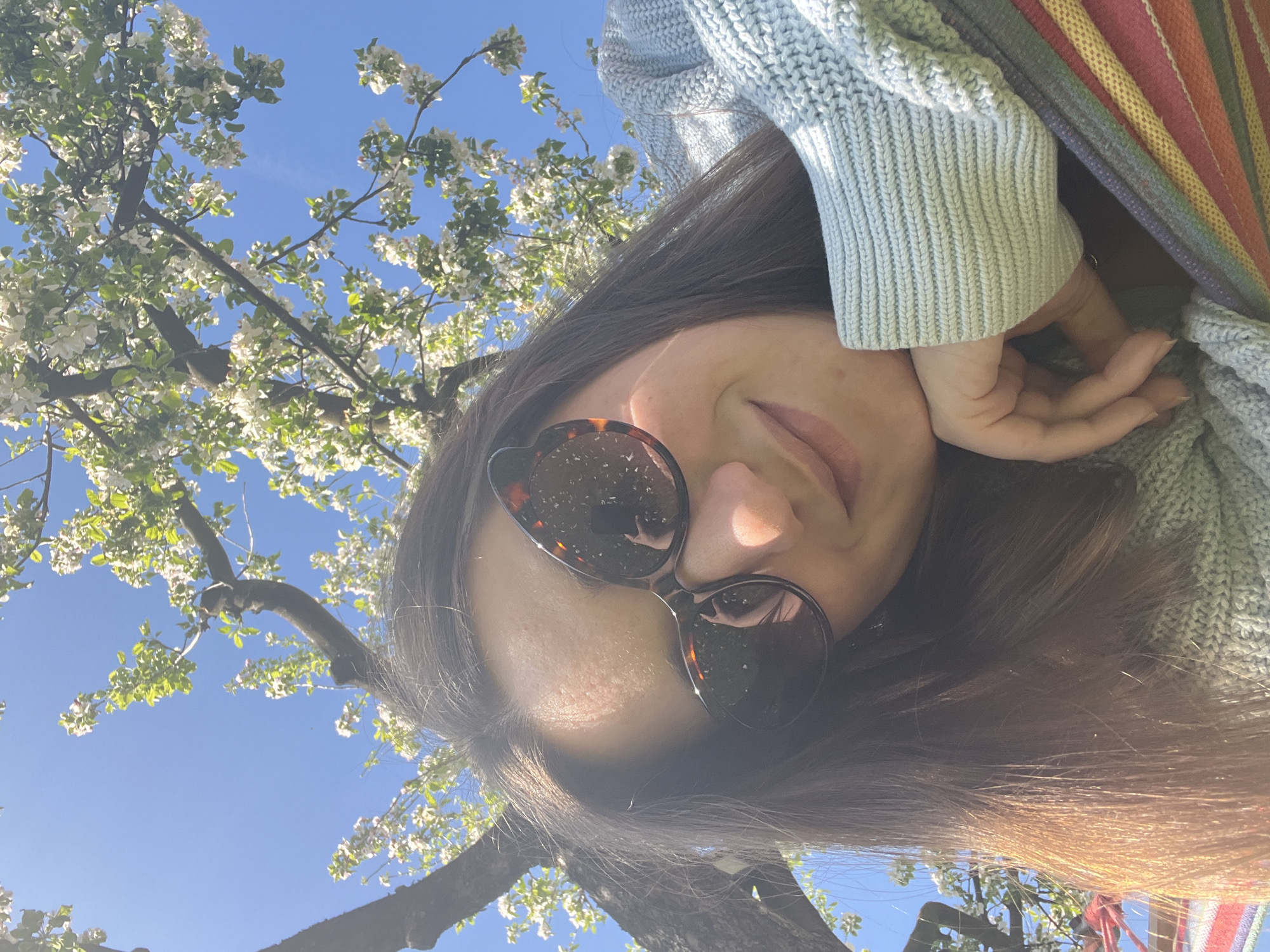 A woman wearing sunglasses, a light blue sweater, and a colorful striped shawl poses for a selfie in Unknown, Unknown. The photo is taken from a low angle, looking up at her face. Behind her, a tree with white blossoms is visible against a clear blue sky. Her hand is resting on her cheek.