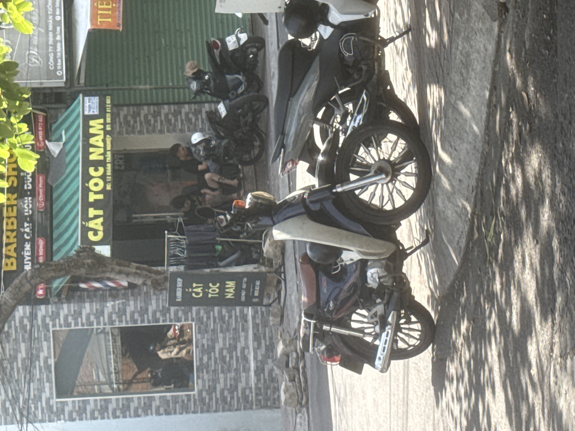 This is a street scene in Bac Nha Trang Ward, Vietnam, featuring a barber shop with the signage "BARBER SHOP CẮT TÓC NAM". The barber shop has a distinctive brick facade and a green awning. In front of the shop, two motorcycles are parked. The motorcycle in the foreground is a dark blue and chrome model with a red seat. The motorcycle behind it is black. Several people are visible sitting on the steps of the barber shop, appearing to be patrons or employees. The lighting suggests it is daytime, with shadows cast from trees.