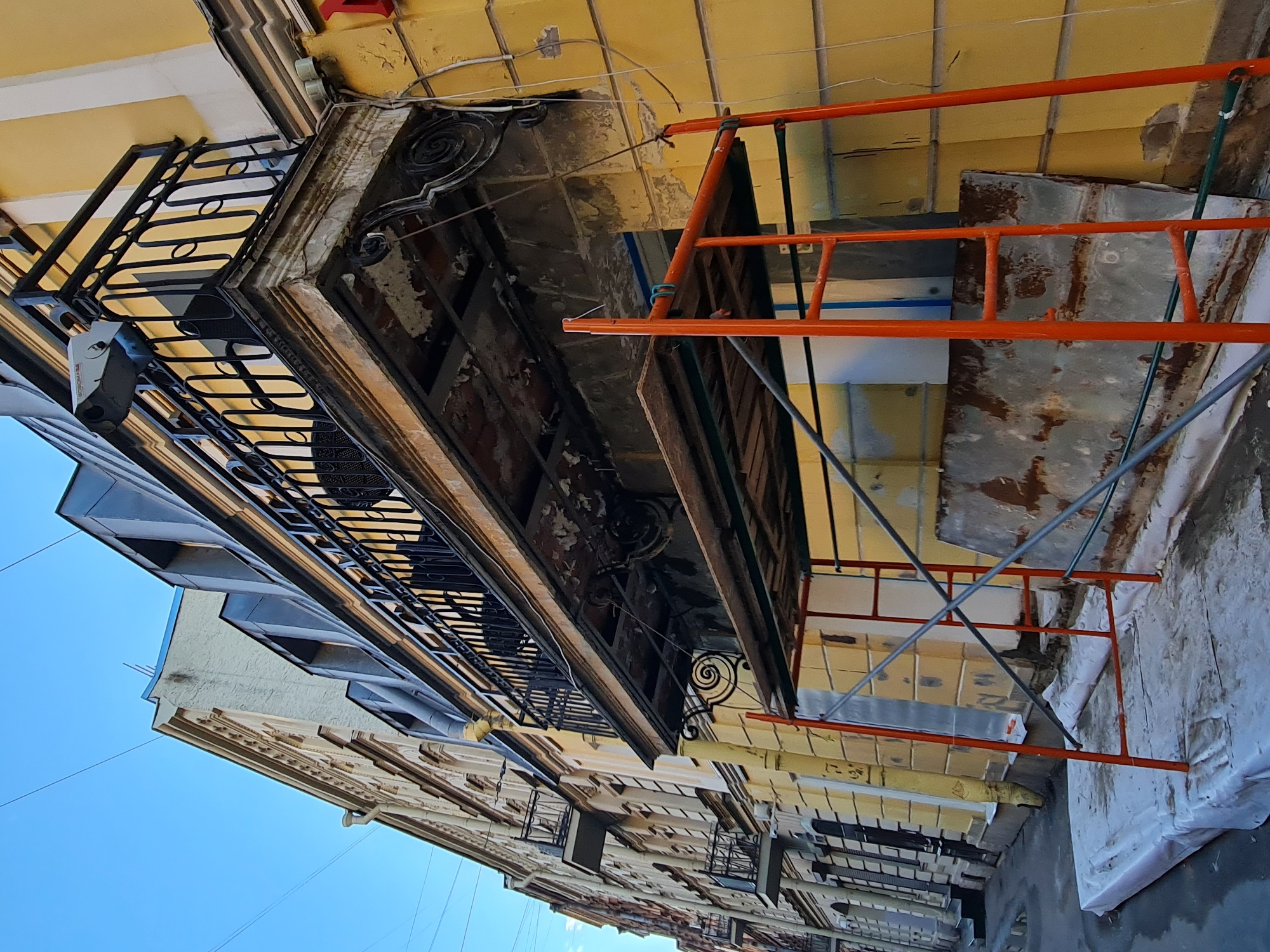 This image depicts a narrow street in Saint Petersburg, Russia, lined with historical buildings. The focus is on the facade of a yellow building undergoing renovation, evidenced by orange scaffolding and protective sheeting covering a large mattress or padding on the ground.
A balcony on the second floor of the yellow building is in disrepair, with peeling paint and exposed brickwork. A black wrought-iron railing adorns the balcony, and a security camera is mounted on its exterior. Above the balcony, dormer windows with dark metal roofs are visible on the building's upper story.
To the left of the yellow building, a row of similarly constructed historical buildings recedes into the distance. These buildings also feature balconies and ornate architectural details. The street itself is wet, suggesting recent rain, and the sky above is a clear blue. The scene captures a moment of urban renewal and preservation within the architectural landscape of Saint Petersburg.