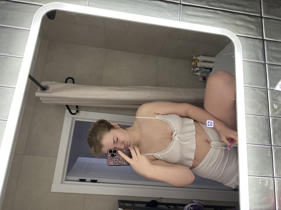 A young woman in a white, frilly two-piece outfit poses in a bathroom mirror, holding a smartphone with a patterned case. Her face is close to the phone, possibly taking a selfie. A shower curtain hangs behind her, and shelves with various bathroom items are visible above. The bathroom features gray tiles. A light-up button is on the mirror.