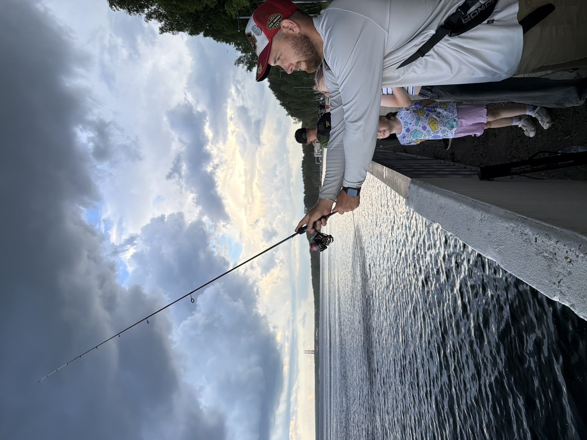 A man is fishing from a concrete embankment next to a large body of water. He is wearing a long-sleeved shirt, a baseball cap, and shorts. He holds a fishing rod with a reel in his hands, casting it into the water. Several other people are visible in the background, including a young girl standing near the man. The sky is cloudy with patches of sunlight breaking through, suggesting either dawn or dusk. In the distance, industrial structures, possibly power line towers, are visible across the water. The location is the embankment of the Votkinsk Reservoir in the city of Chaikovsky, Russia.