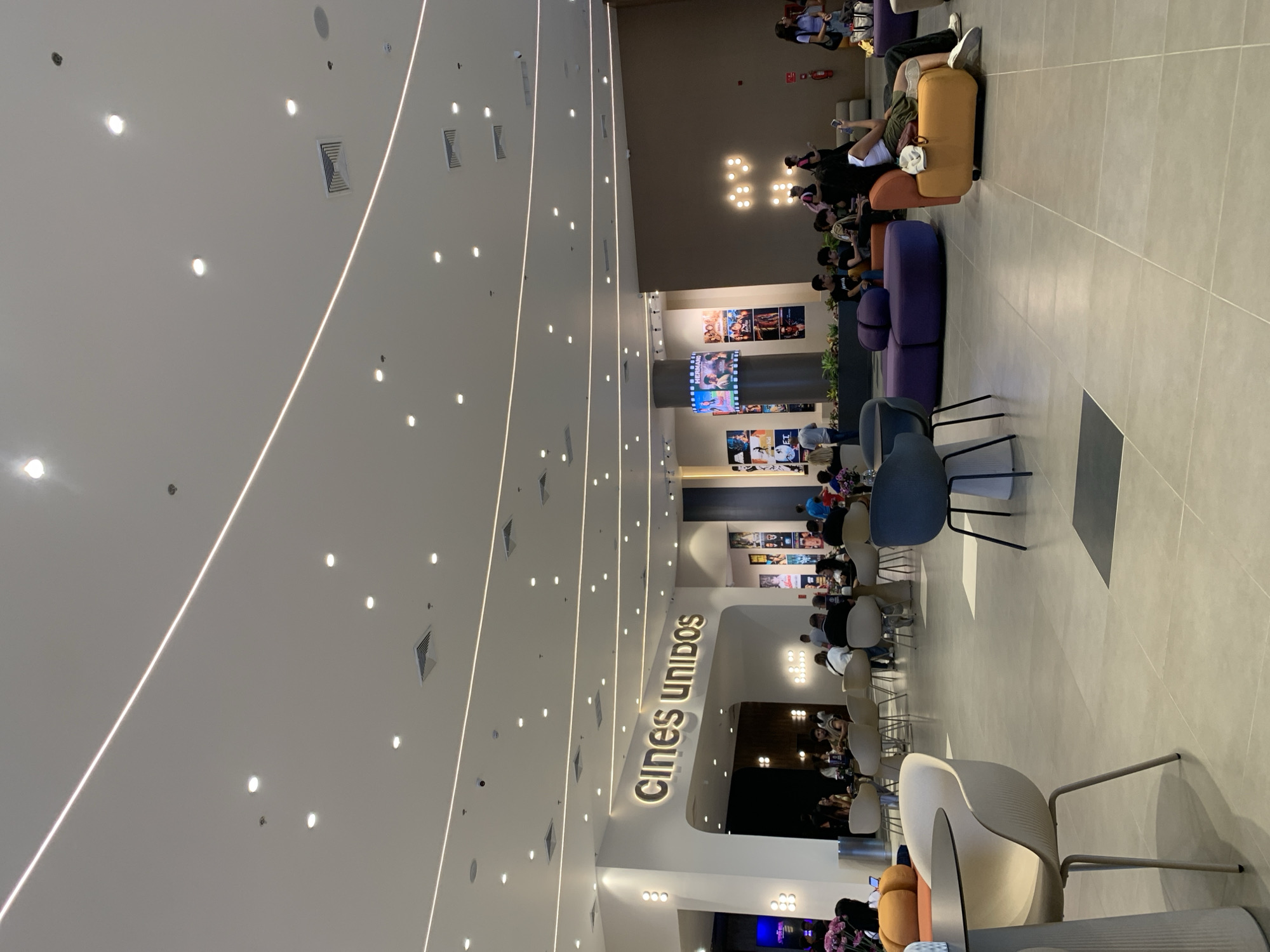 The image depicts the interior of the Cines Unidos movie theater in Porlamar, Venezuela. The scene includes seating areas with colorful lounge chairs and tables, interspersed with rows of chairs. Patrons are visible, sitting and socializing. Movie posters and digital screens adorn the walls. The theater's high ceiling is characterized by linear lighting and recessed lights. The floor is tiled. The scene suggests a pre-movie or intermission period.