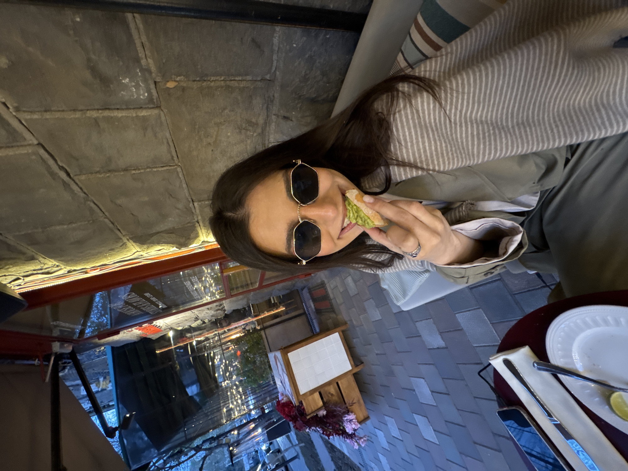 The image depicts a woman with long brown hair and sunglasses seated outdoors at a restaurant, possibly in Yerevan, Armenia. She is eating a sandwich. A table with cutlery and a plate is visible, along with a glimpse of the city's exterior. The setting includes a brick-like ground and outdoor decorations like lights and plants.