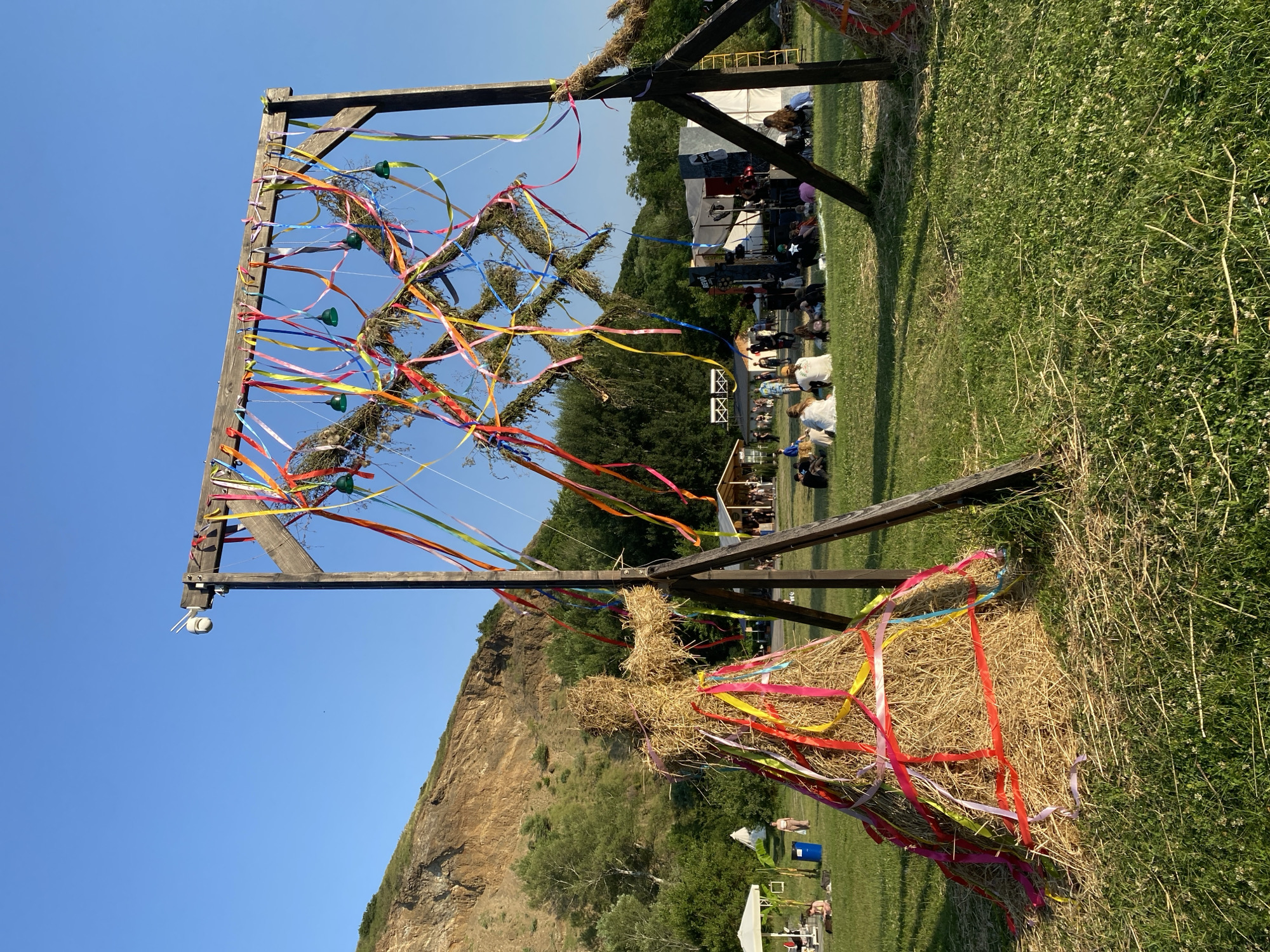 In Asureti, Georgia, an outdoor festival takes place on a grassy field with a wooded hillside in the background. A large wooden structure adorned with colorful ribbons and greenery stands prominently, while a scarecrow figure made of hay, also decorated with ribbons, is situated to its left. In the distance, a stage with lighting and speakers is visible, with a crowd gathered in front of it. Several tents and other structures are scattered across the field, and people are seen sitting on the grass, enjoying the event. The sky is clear and blue.