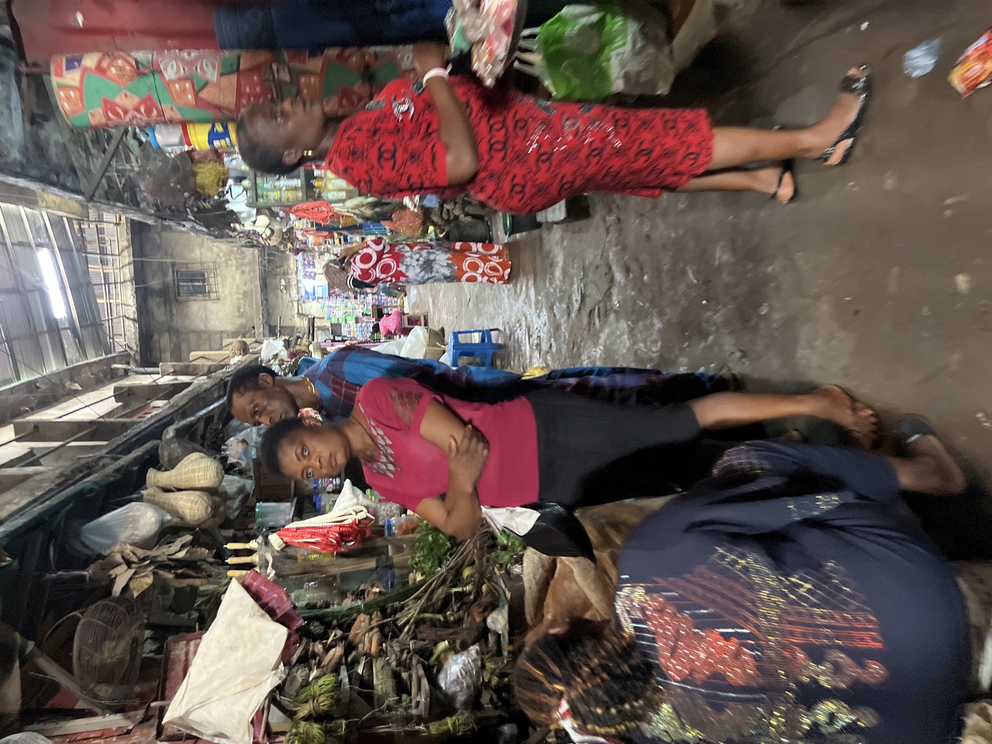 The image depicts an indoor market scene in Nkpor, Nigeria, likely during daylight hours, though the lighting is dim and artificial. Several people are present, some appearing to be vendors or shoppers. In the foreground, a woman in a bright pink shirt and black pants is looking towards the camera, her arms crossed. Behind her, a man in a blue checkered shirt is leaning forward, also looking towards the camera. Further back, another woman is lying down or resting, wearing a red dress with a repeating pattern. Another person in a red and white patterned dress is visible. The market stalls are filled with a variety of goods, including woven baskets, bags, what appear to be food items like leafy greens and roots, and possibly household supplies. The ground is muddy and uneven, indicating a rustic and potentially damp environment. The overall atmosphere seems somewhat subdued, with the individuals in the foreground appearing serious or contemplative. No discernible text is visible in the image.