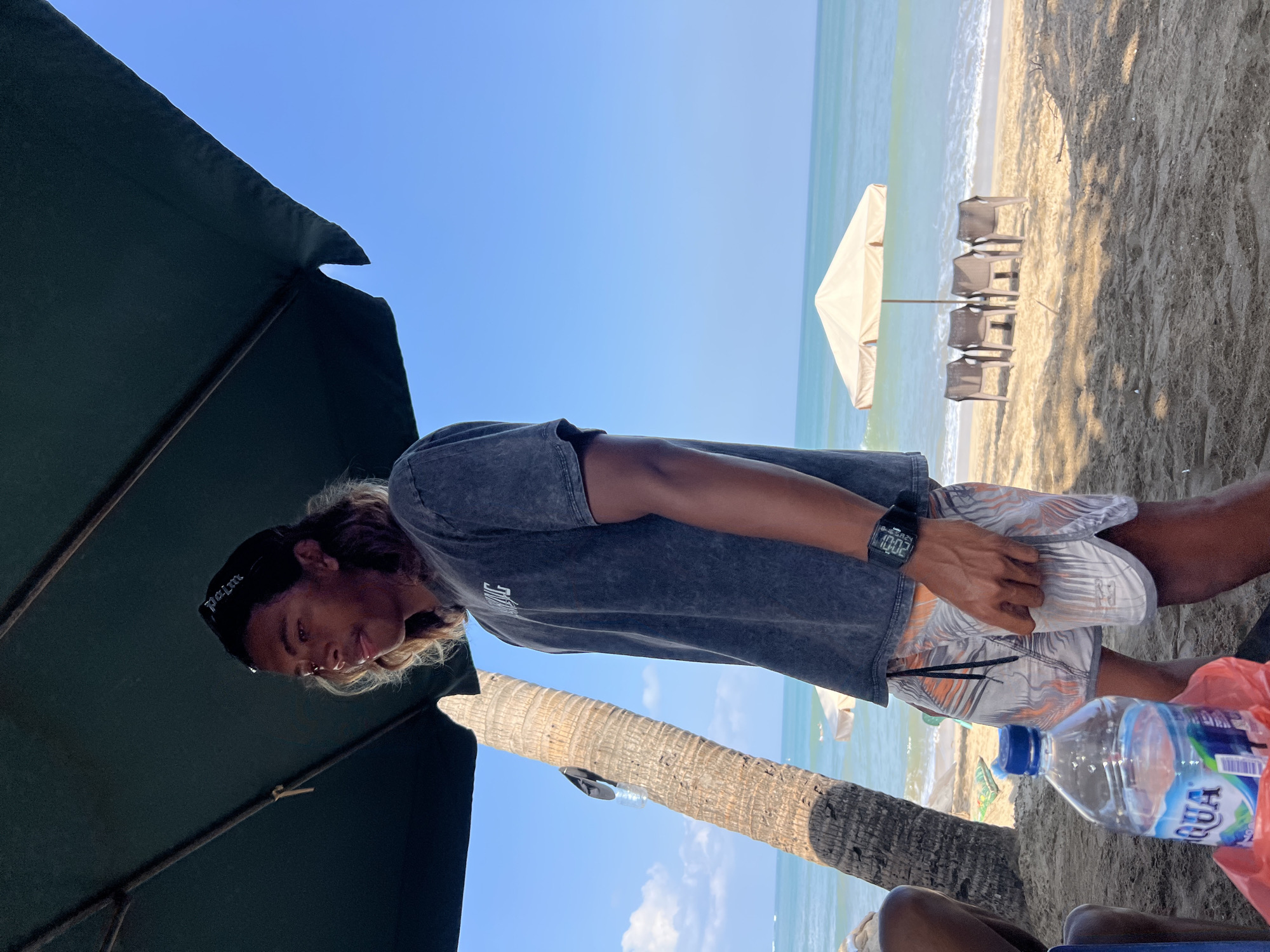 A medium shot shows a young man standing on a beach in Kuta, Indonesia. The man has long, wavy brown hair and is wearing a grey t-shirt, patterned shorts, and a black baseball cap. He is standing under a large green umbrella, with a palm tree to his left. In the foreground, a bottle of Aqua brand water sits on the sand. In the background, the ocean is visible, with a few beach chairs and umbrellas on the sand. The sky is clear and blue.