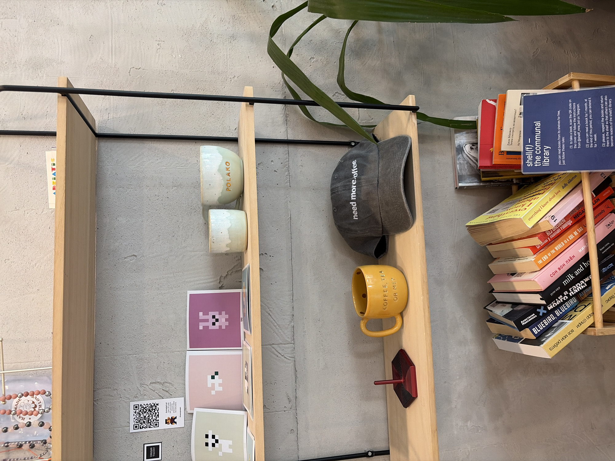 The image shows a multi-tiered shelf in what appears to be a retail space or cafe, likely in Budva, Montenegro. The shelves are constructed of light-colored wood with black metal framing.
On the top shelf, there are decorative beaded necklaces and small printed cards. Below this, on the second shelf, are three small pixelated art prints featuring animal-like characters, two ceramic cups, one with "POLAKO" written on it.
The third shelf holds a bright yellow mug with the text "COFFEE, TEA OR ME?" embossed on it, a small red geometric object, and a grey baseball cap with "need more coffee" embroidered in white. A portion of a green plant is visible to the right of this shelf.
The bottom shelf displays a stack of books leaning in a wooden crate. A blue sign with white text is propped up in front of the books, detailing the rules for "shell(f) - the communal library." The text instructs users to scan a QR code on book covers to borrow and extend book loans. The background is a textured concrete wall.