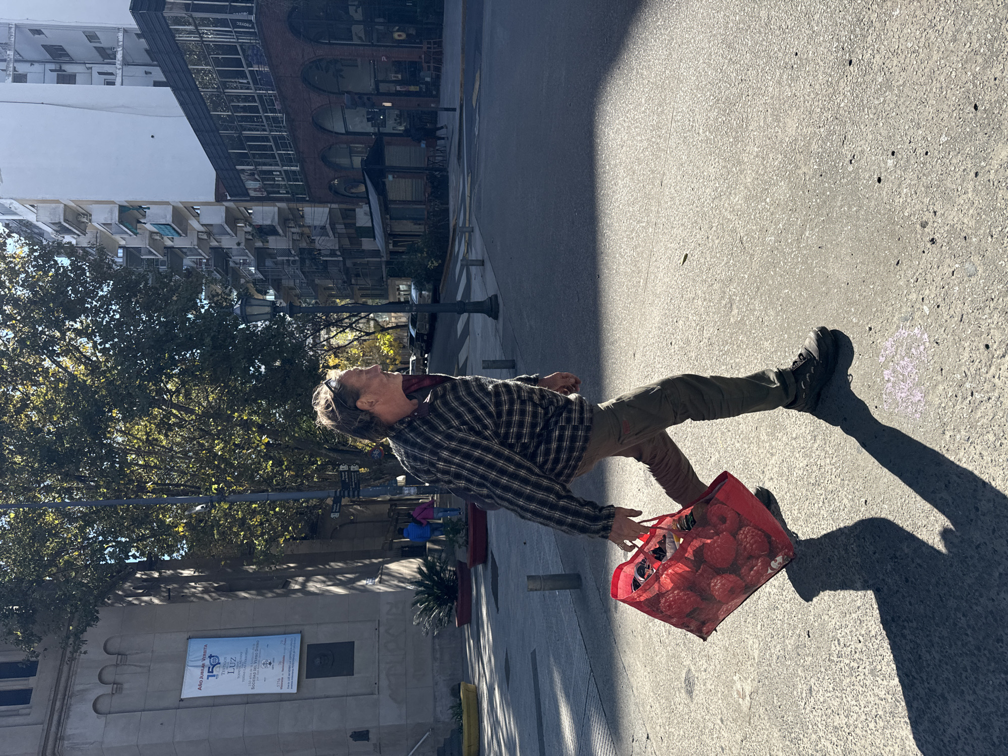 A medium shot depicts a man on a city street. He's wearing a plaid shirt, khaki pants, and boots. He's carrying a red mesh bag filled with red items. The street is concrete and is partially shaded. Buildings and trees are visible in the background. A banner, advertising the Society of the Divine Word, is attached to one of the buildings. The man is in Buenos Aires, Argentina.