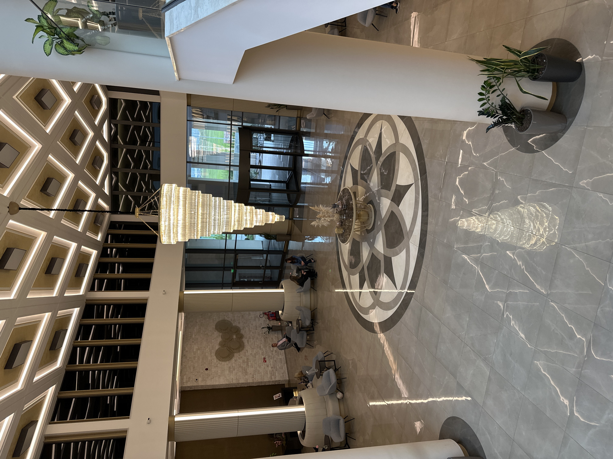 The image depicts the interior of a large lobby, possibly a hotel, in Kazanlak, Bulgaria. The floor is covered with polished gray marble tiles with a circular design in the center featuring a star pattern. A multi-tiered crystal chandelier hangs from the ceiling. Seating areas with armchairs and sofas are arranged around the central design. Several people are sitting in the seating areas. There are large planters with greenery near a large pillar on the right. Large windows are visible in the background. The architectural style is modern, with clean lines and geometric patterns. The building features a lighted diamond pattern architectural design.