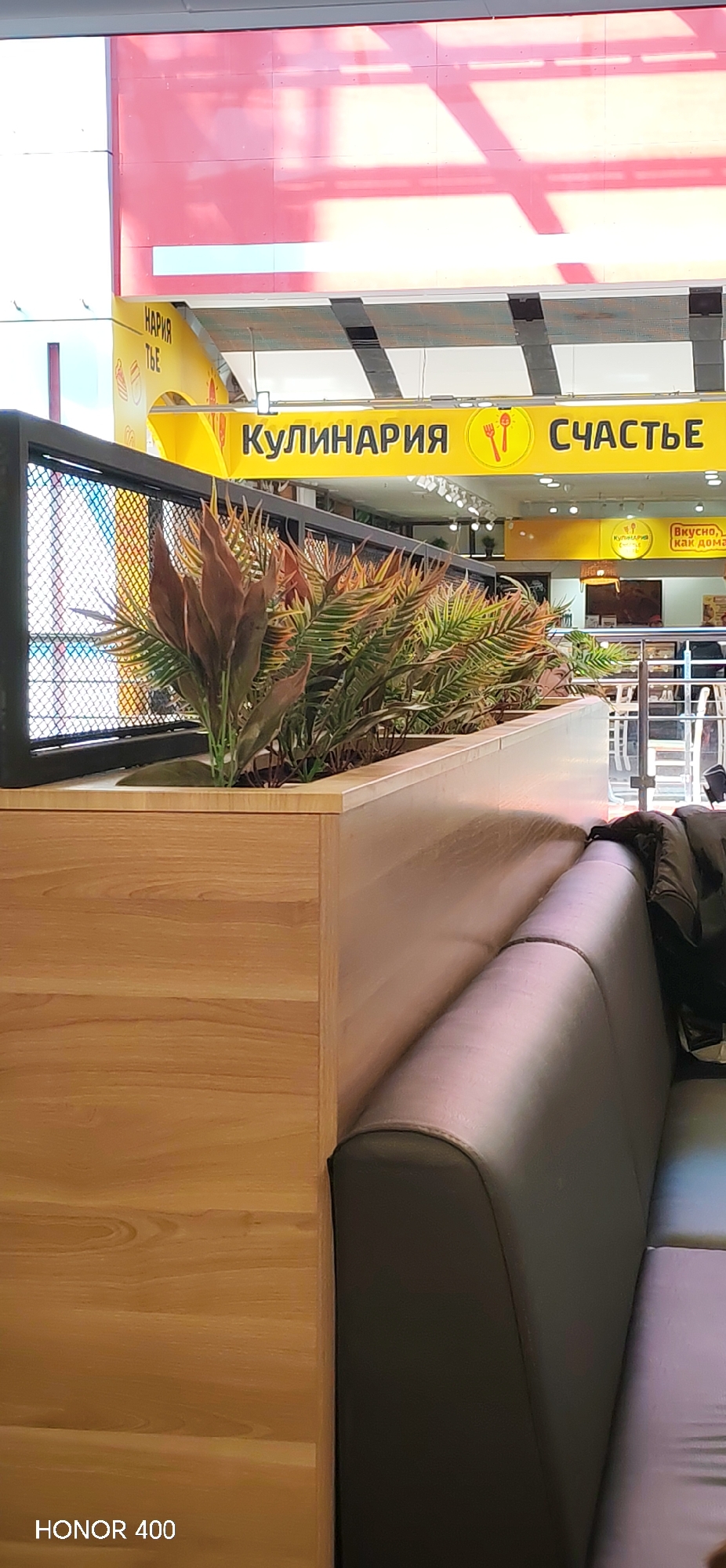 The image shows the interior of a food establishment in Izhevsk, Russia. A sign reads "КУЛИНАРИЯ СЧАСТЬЕ," which translates to "CULINARY HAPPINESS." There is a planter box with plants and a black metal fence on top. Adjacent to the planter is a dark gray leather booth. A jacket is placed on the booth's corner. The floor appears to be light-colored wood. The text "HONOR 400" is displayed in the lower left corner of the image.
