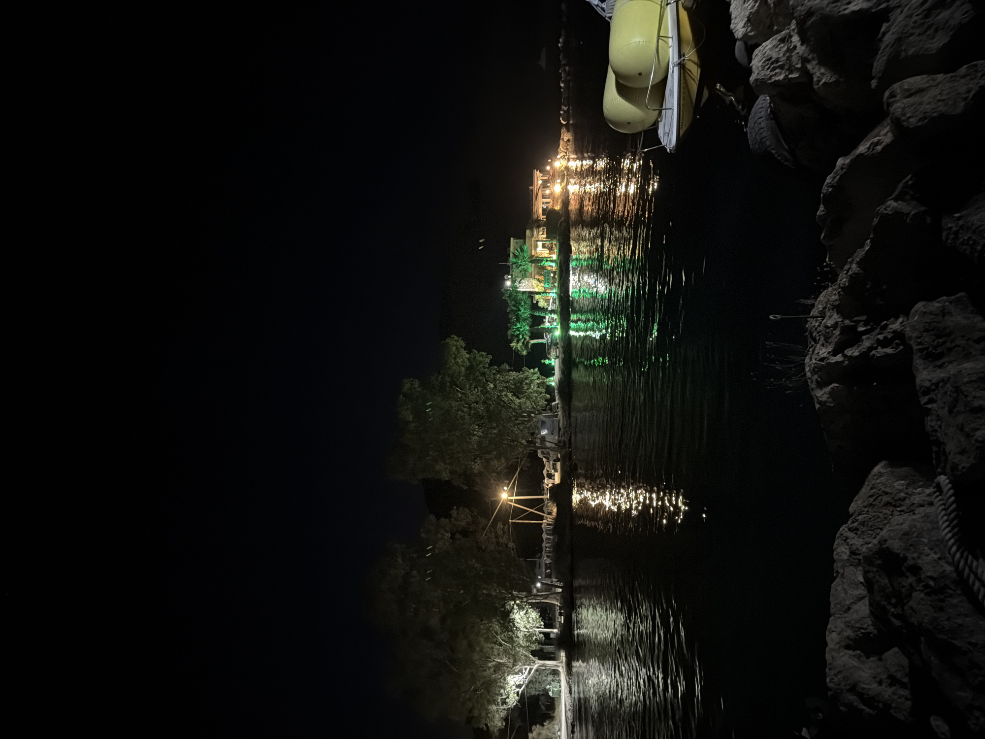 The image captures a nighttime scene at a waterfront establishment in Georgioupoli, Greece. A body of water, reflecting the lights from the shore, occupies the lower half of the frame. On the left bank, trees are illuminated by artificial lights, casting reflections on the water. Further along the shore, a building is brightly lit with green and yellow lights, suggesting it is a restaurant or entertainment venue. Palm trees are visible in front of the building, also illuminated. In the foreground on the right, a yellow paddle boat is partially visible, docked or moored near a rocky shoreline. The sky is completely dark, indicating it is well past sunset. There are no people clearly visible in the image.