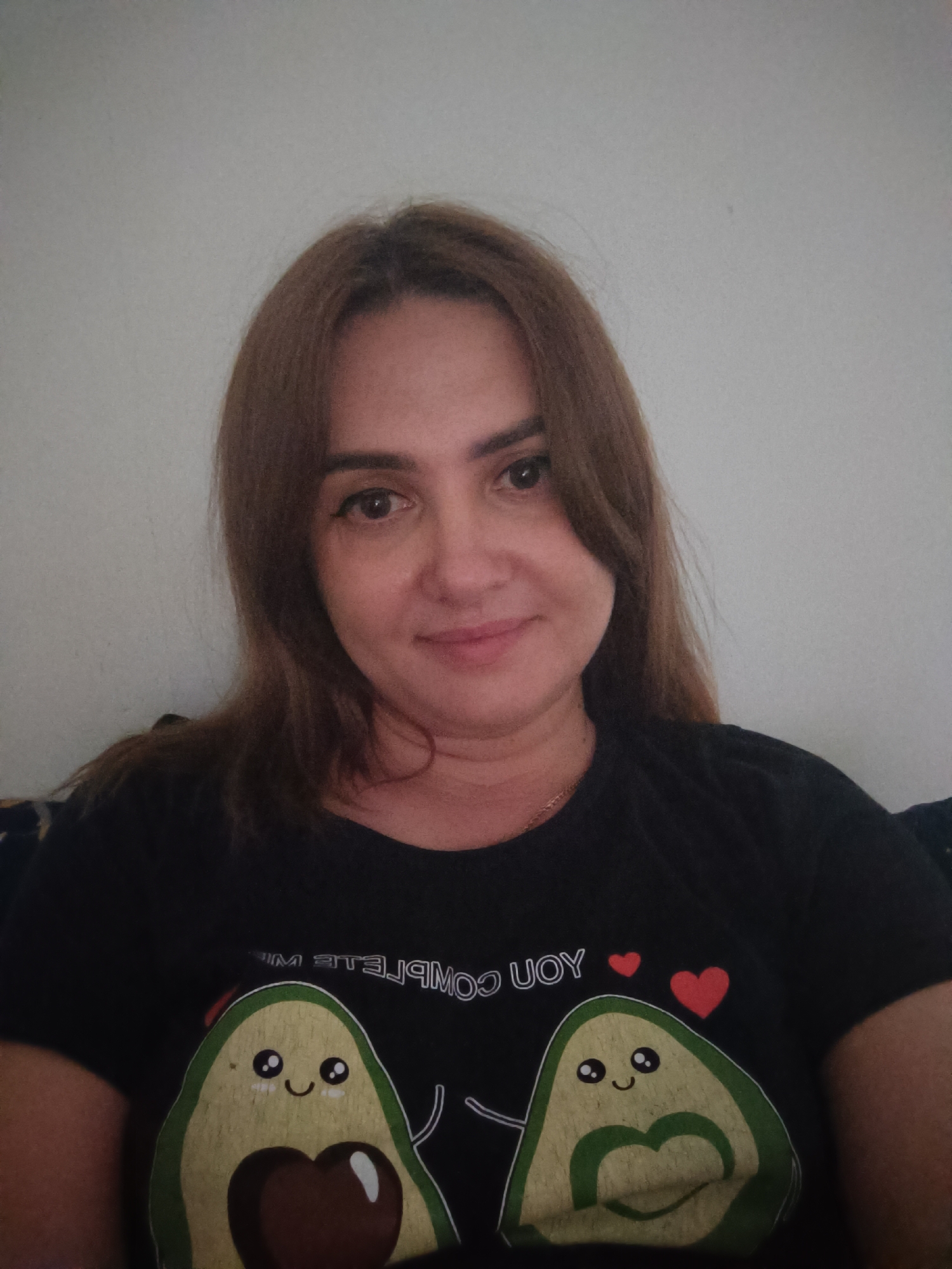 The image is a medium shot selfie of a woman with brown hair, fair skin, and brown eyes. She is wearing a black t-shirt with a graphic design featuring two avocados, hearts, and text. The background is a plain, light-colored wall. The woman is looking at the camera with a slight smile. There are no discernible landmarks or indications of a specific city or location within the image itself.