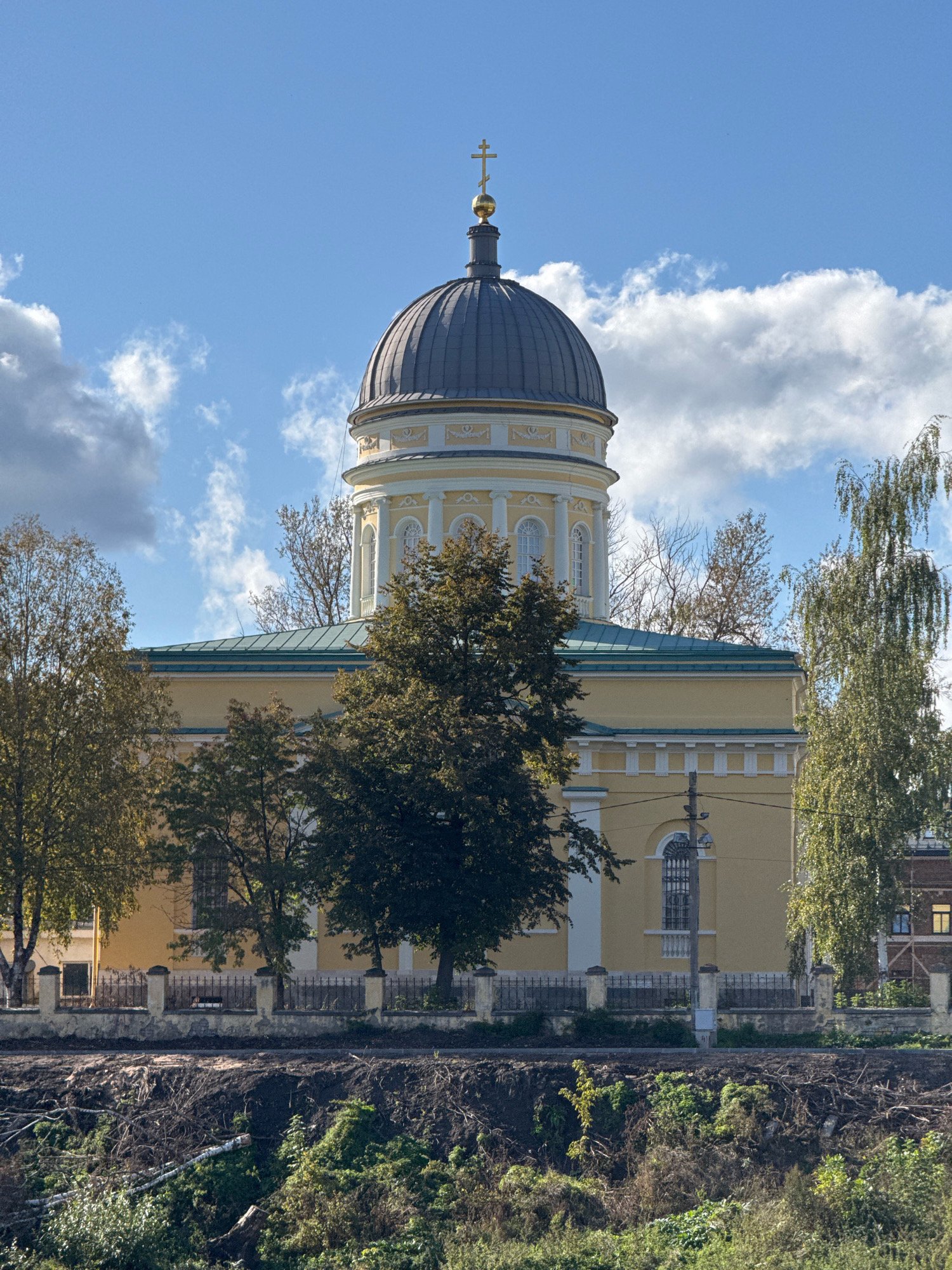 The image shows a large, yellow, neoclassical church with a dark dome topped by a golden cross, situated in Torzhok, Russia. The church features an octagonal drum with arched windows and decorative columns. A green roof runs along the main body of the building, which is partially obscured by trees. A black metal fence with stone pillars separates the church grounds from a strip of barren, disturbed earth in the foreground. Beyond the church, a brick building with illuminated windows is visible. The sky above is blue with scattered white and grey clouds. The scene appears to be set in the daytime.