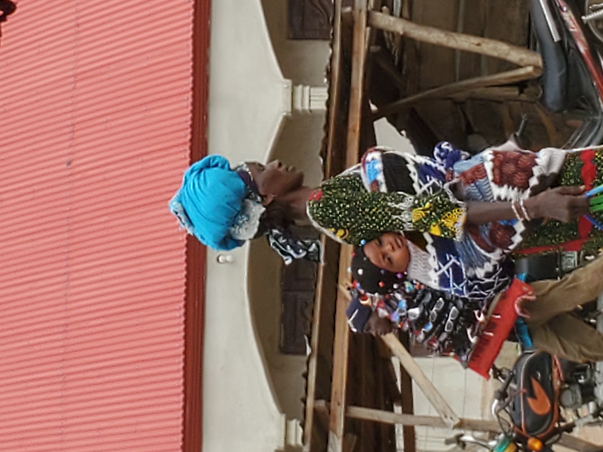 This vertically oriented image captures a bustling outdoor scene, likely a street or market in Mararaba, Nigeria, during daylight hours.
The foreground is dominated by three individuals. A woman with dark skin, wearing a vibrant blue headwrap and a patterned green, yellow, and black dress, stands prominently on the left, looking upwards. In front of her, a child with a darker complexion and a patterned head covering looks directly towards the viewer with a neutral expression. Another individual, partially obscured, is visible behind the child.
The scene suggests a commercial setting. Numerous pairs of sunglasses are displayed on racks around the child and the third person, indicating a vendor's stall. A hand adorned with white bracelets holds several colorful stick-like objects, possibly for sale.
In the background, a tall red corrugated metal structure forms a prominent wall or roof on the left. Behind the people, light-colored buildings with distinct architectural features and wooden support beams suggest a semi-permanent or informal market structure. Towards the lower right, parts of a motorcycle or bicycle are visible, including handlebars and a black bag with a distinct orange logo attached to it. A red object, possibly a small keyboard or display stand, is also present near the bottom. No discernible text is visible in the image.