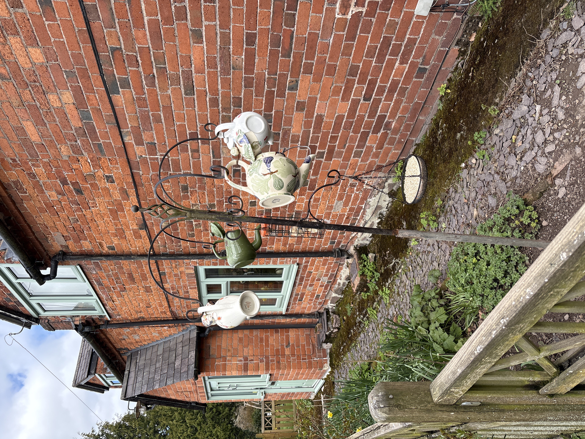 A sideways-oriented outdoor shot depicts a brick building with light green window frames in Shrewsbury, United Kingdom. A wrought iron stand is positioned in the garden area alongside the building. The stand has several ceramic teapots and teacups hanging from it. A bird feeder is attached to the stand, filled with seeds. The ground is covered with small stones, moss, and various plants. A weathered wooden fence is in the foreground.