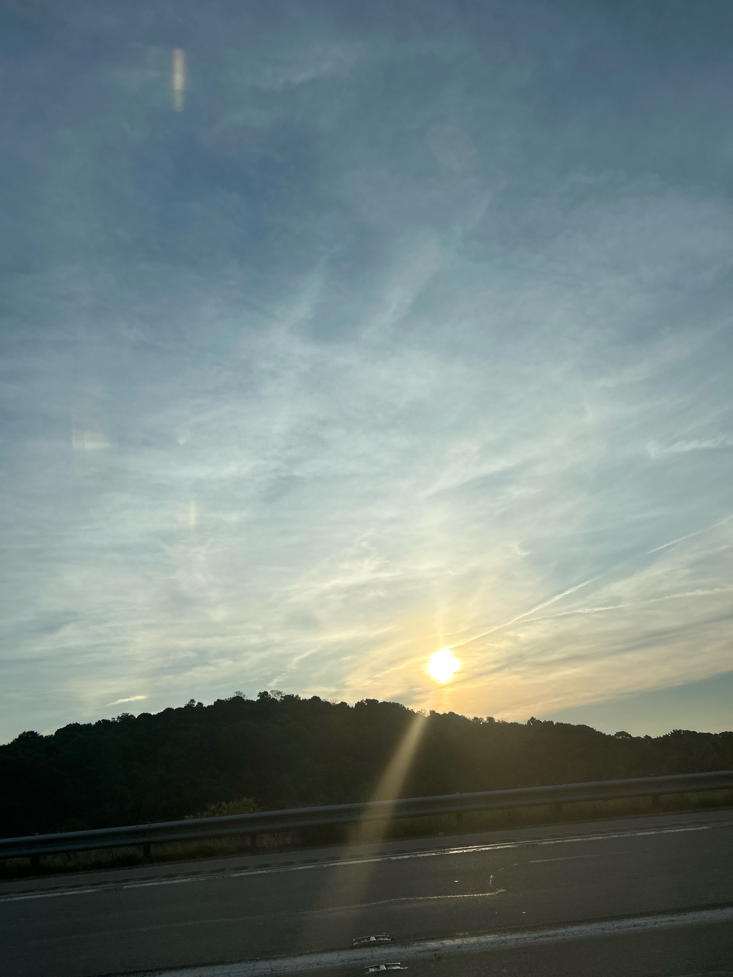 The image captures a scenic view of a hillside covered in dark green trees against a backdrop of a pale blue sky with wispy white clouds. The sun, a bright orb, is positioned just above the treeline, casting a golden glow and long rays of light across the scene. In the foreground, a paved road with white lane markings is visible, separated from the hillside by a metal guardrail. The overall impression is that of a serene landscape, possibly viewed from a vehicle traveling along the road. The location is identified as Robinson Township, United States of America.