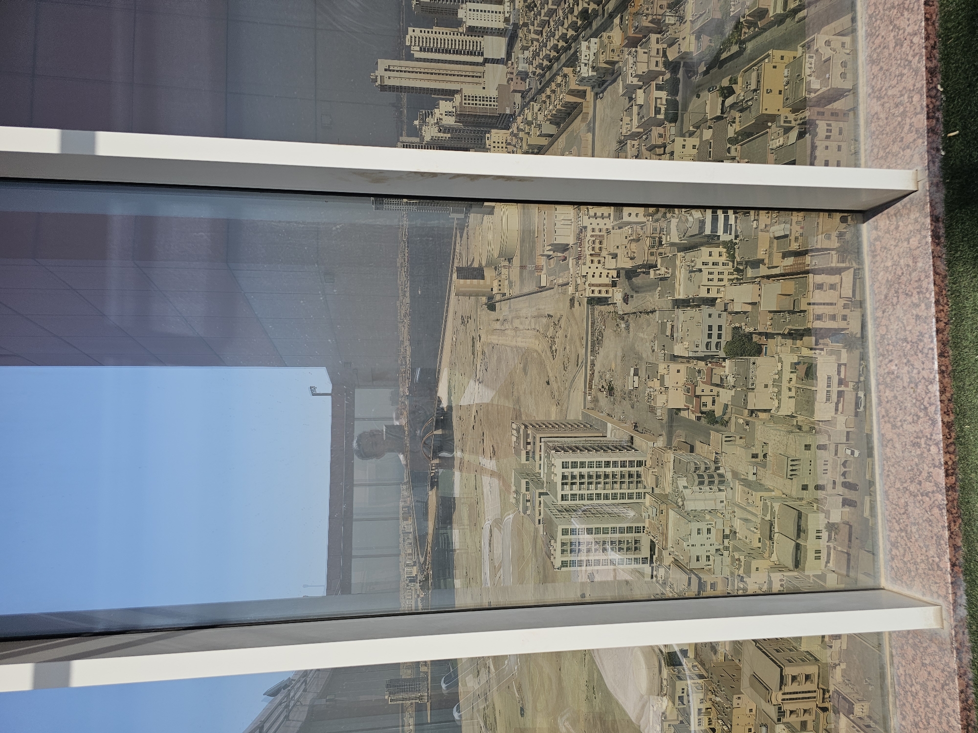 The image shows a reflection on a window of a city with low-rise buildings and some taller structures. The reflection also includes a person's face and upper body. The city appears to be a model or miniature representation, possibly a tourist attraction or an architectural display. The buildings are mostly light-colored and closely packed, with streets visible between them. In the distance, there are more modern high-rise buildings and what looks like a body of water. The sky in the reflection is clear and blue. The location is identified as Manama, Bahrain, suggesting this is a view of a model of the city. The foreground shows a hint of greenery and a textured surface, likely part of the viewing area for the model.