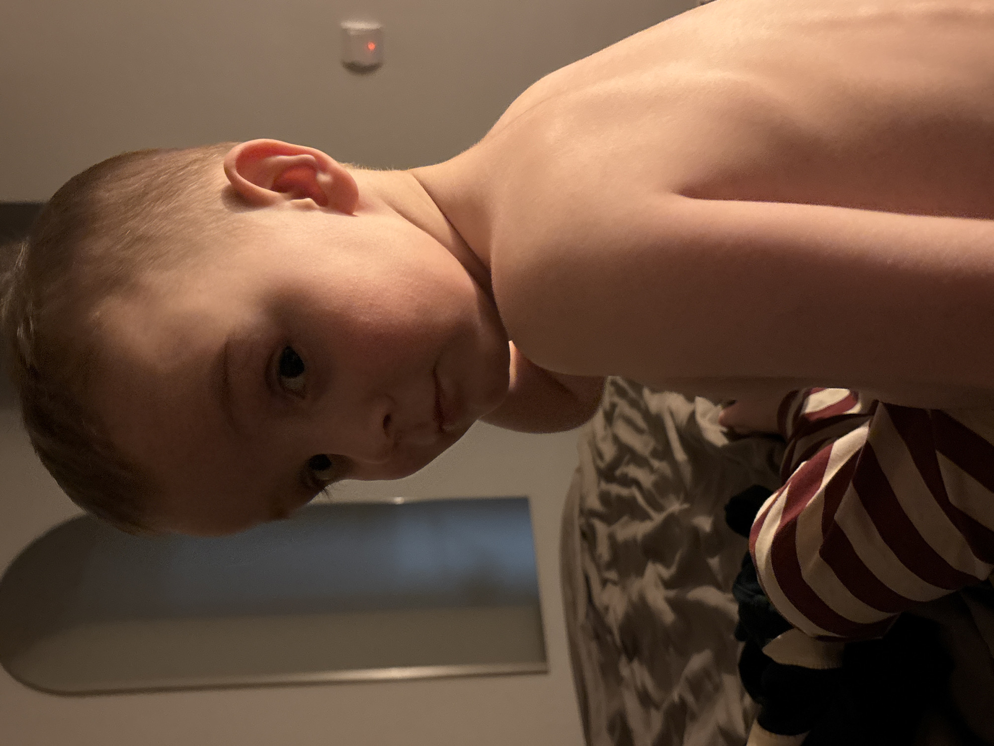 A close-up, medium-lit, tilted indoor shot depicts a shirtless young boy with short blond hair. He is looking down with a slight pout. His ear is visible. Behind him is a grey textured bedspread with a striped white and red fabric on top, and a dark object underneath. In the background, a section of the wall is visible with an arch-shaped window or opening. A small object, possibly a smoke detector, is visible on the ceiling. The location is indeterminable as Chaikovsky, Russia.