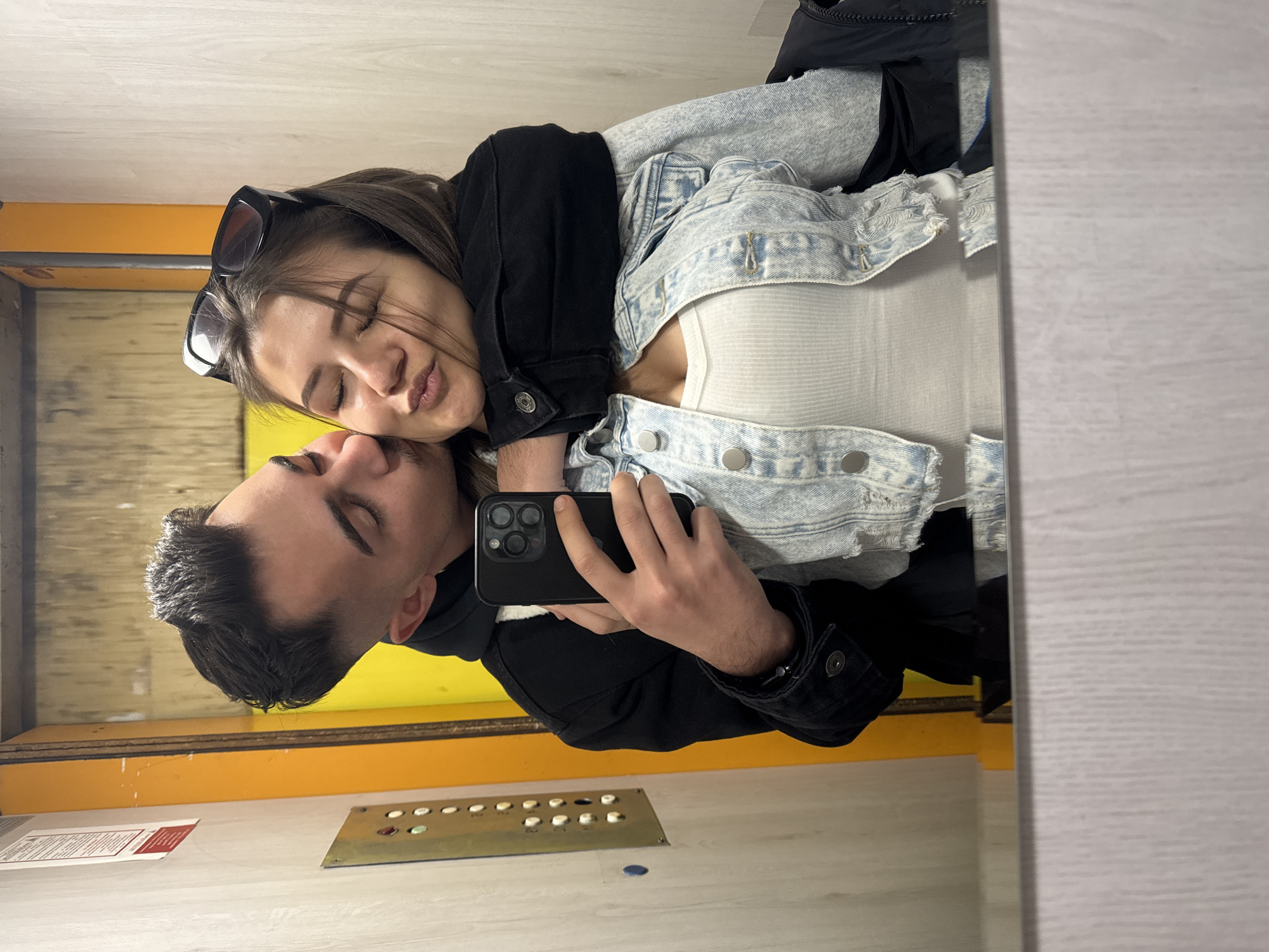 Two people are reflected in a mirror. One person has dark hair, wearing a black jacket over a dark hoodie. The other person has dark hair, wearing sunglasses on their head, a white shirt, and a light denim jacket over a black jacket. One person is holding up a black smartphone. The background includes a yellow wall and elevator buttons and a sign. This photograph appears to be taken in an elevator. There is not enough information to accurately determine the location of Sofia, Bulgaria.