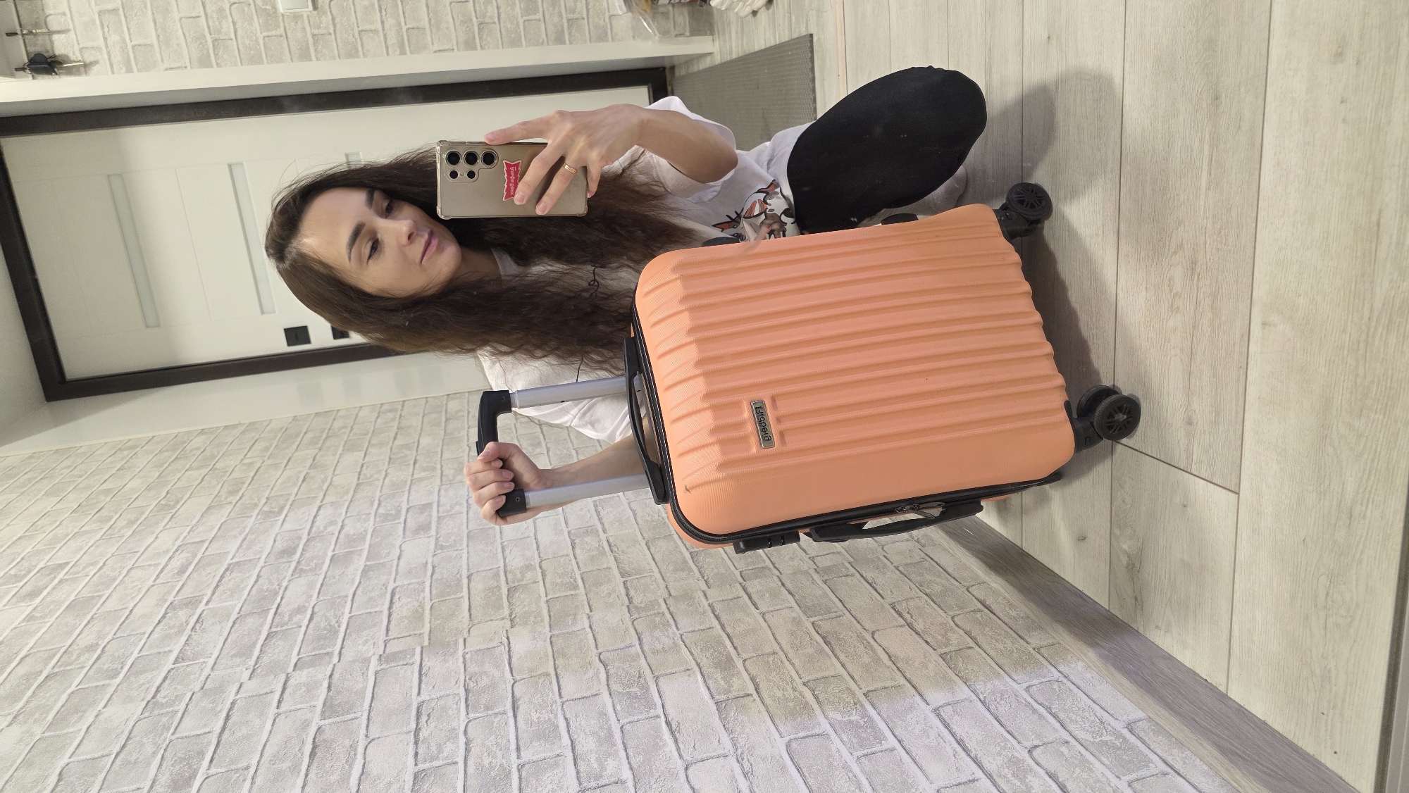 A woman with long, dark, wavy hair is kneeling on a light-colored wooden floor, holding the handle of an orange, ribbed suitcase. She is wearing a white t-shirt with a graphic print and black pants. She is looking at her gold smartphone, which she holds in her right hand, and appears to be taking a selfie. The suitcase has black wheels and a black retractable handle. Behind the woman is a white brick wall. To her right is a doorway with a dark frame and a white door. The scene appears to be indoors, possibly in a hallway or near an entrance. The image does not provide any specific clues to pinpoint a location in Bulgakovo, Russia, or any other city.