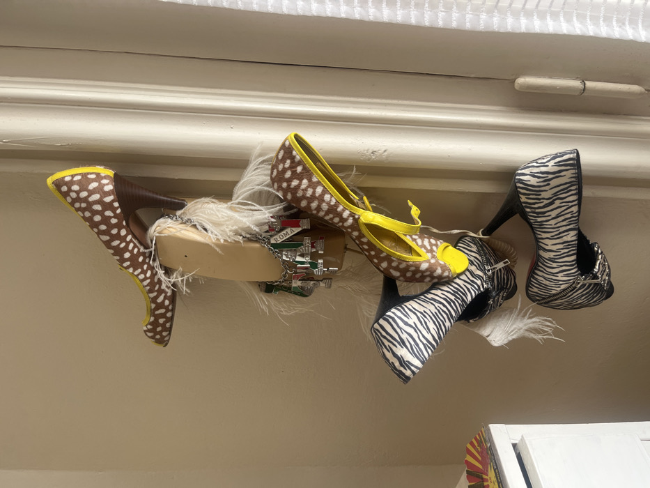 The image depicts an indoor scene focused on a vertical display of high-heeled shoes and decorative items mounted on a wall. No individuals are present.
Four high-heeled shoes are arranged on a light-colored, rectangular block attached to a beige-tan wall. The topmost visible shoe is a brown and white spotted (deer print) peep-toe pump with a yellow trim and a brown heel, oriented horizontally to the right. Below this, a brown and white spotted high-heeled sandal with yellow straps hangs downwards. The two lowest visible shoes are black and white zebra-print high heels, both featuring black heels and zipper accents, hanging vertically downwards. White feathers are interspersed around the central block, particularly between the shoes. Draped over the middle section of the block is a silver-toned charm bracelet, adorned with multiple charms, several of which display the Italian flag colors (green, white, red) and the text "ROMA."
The background consists of a plain, light beige-tan wall. To the right, a white architectural element, consistent with a door or window frame featuring vertical moldings, extends from top to bottom. A sheer white curtain or blind is partially visible on the far right. In the lower-left corner, a segment of white furniture, possibly a cabinet, is visible, topped by a fan-shaped object displaying red and yellow hues. The scene is illuminated by ambient light. No specific location within Buenos Aires, Argentina, can be determined from the visual information provided.