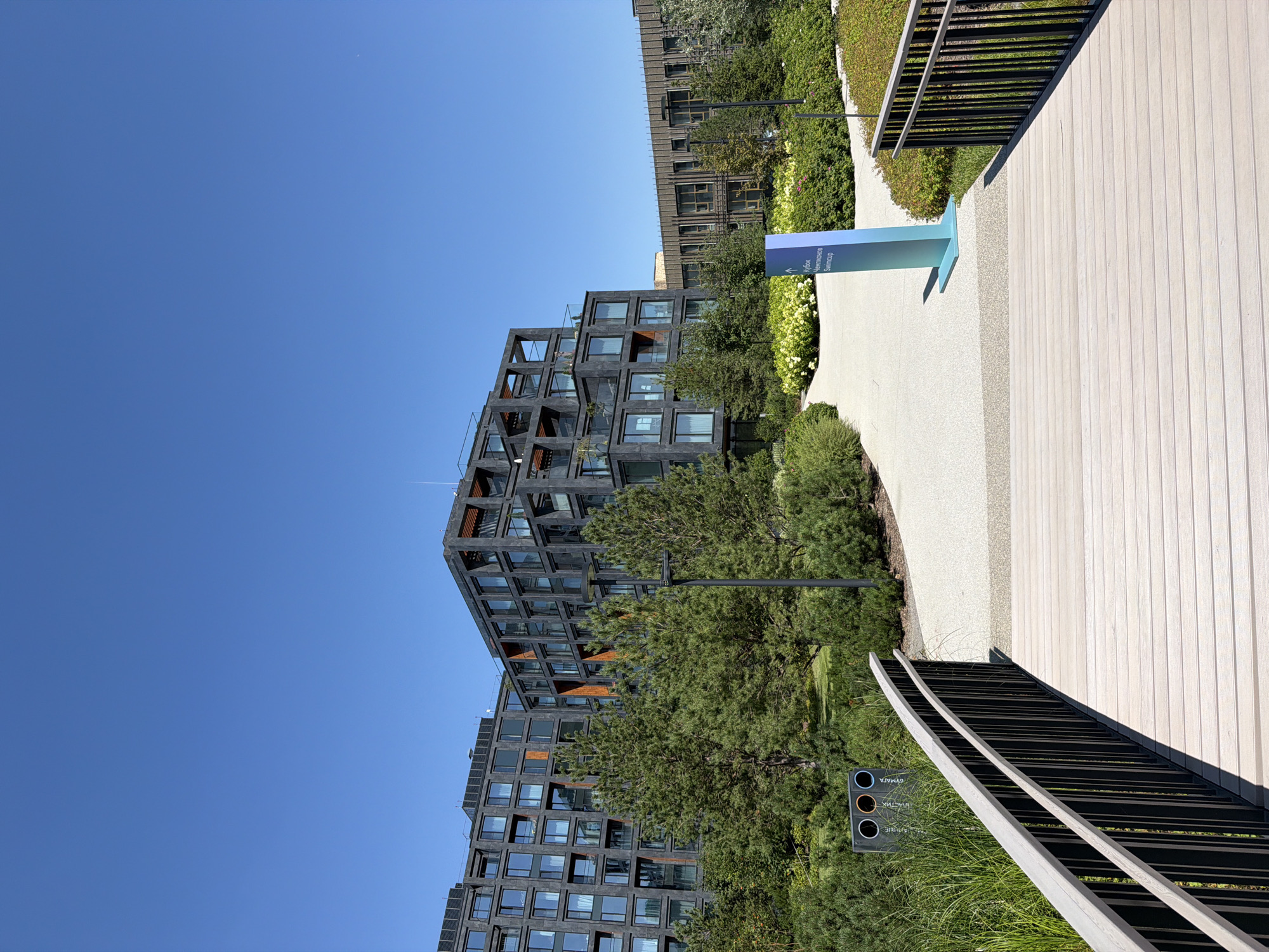 The image depicts the exterior of the Skyport apartments in Moscow, Russia. The building, characterized by its dark, modern facade and geometric design, is situated amidst lush greenery. A wooden bridge with black railings spans a path, leading towards the building. In the foreground, the bridge's textured wooden planks are visible, illuminated by sunlight. To the left of the bridge, a green trash receptacle with separate compartments for recycling is present. A tall, teal-colored sign with text in Russian points towards "Кубок Чечнинов" (Chechninov Cup) and "Swimcup". The surrounding landscape is a manicured garden with various trees and shrubs, creating a serene and contemporary urban environment. The sky above is a clear, bright blue.