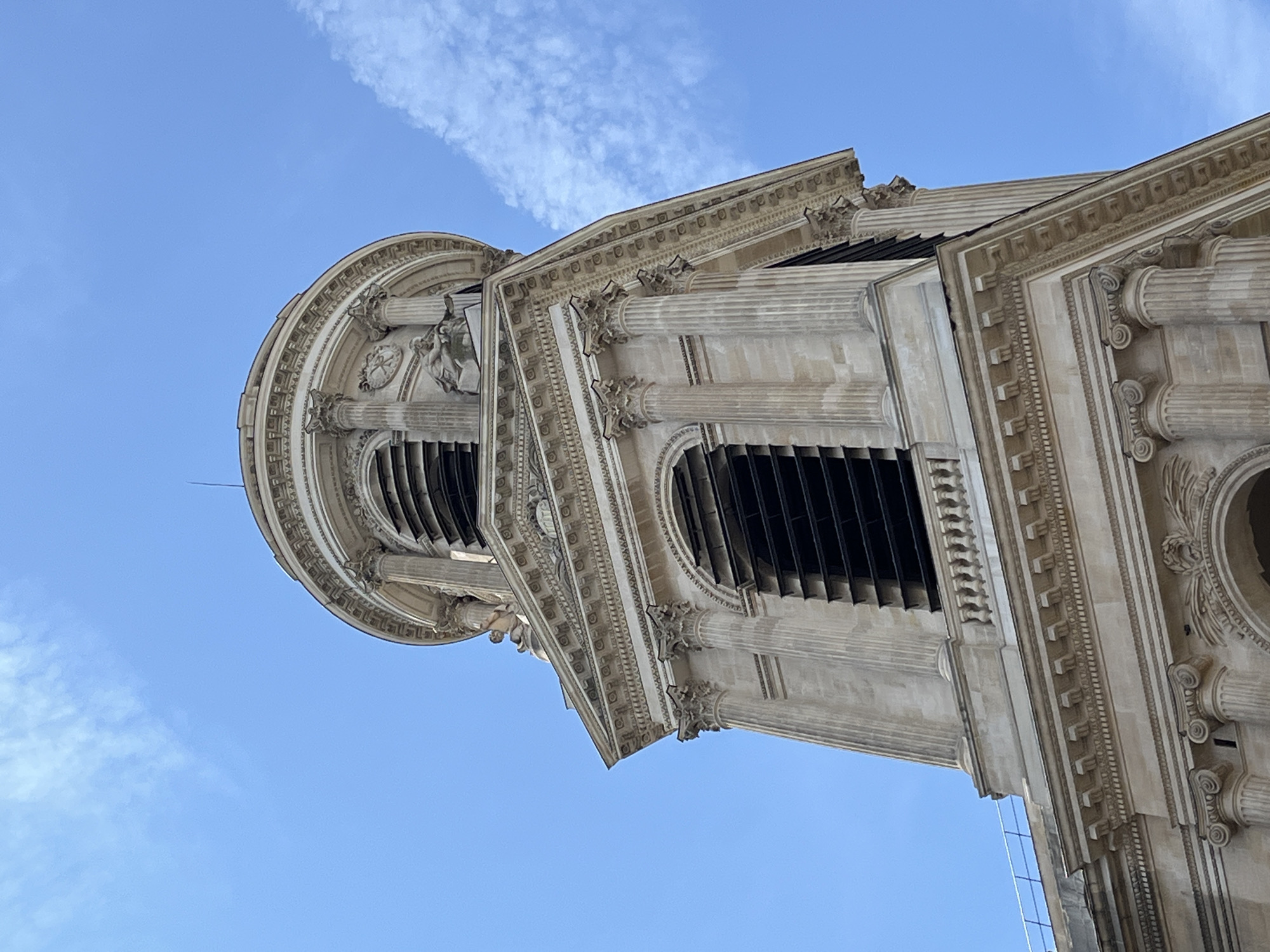 A low-angle, full shot captures the top of a pale stone bell tower against a bright blue sky with a few wispy white clouds. The tower exhibits classical architectural features, including ornate carvings, columns with Corinthian capitals, and arched openings with dark louvers. A circular section at the very top features a dome-like structure with more columns and decorative sculptures. The stone appears weathered but intact. The image is cropped to focus on the upper portion of the structure, implying it is part of a larger building, likely a church or cathedral, within the city of Paris, France. No people or specific actions are depicted.