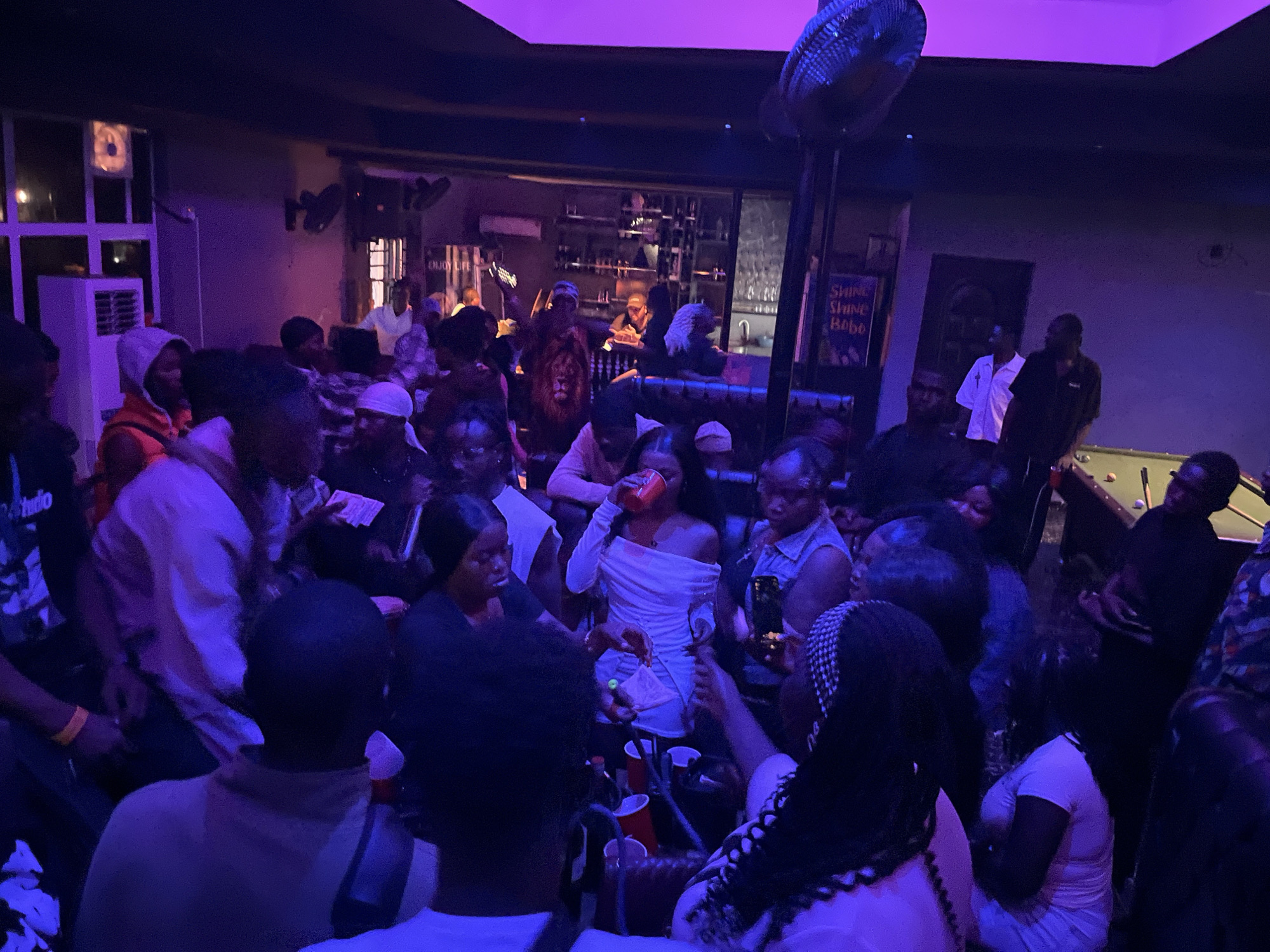 In the city of Unknown, Unknown, an indoor social venue, likely a bar or lounge, is depicted as densely occupied by approximately 30-40 individuals. The scene is illuminated by dim, pervasive purple artificial lighting. Most occupants appear to be young to middle-aged adults.
On the left side of the background, multi-paned windows are visible above a white, wall-mounted air conditioning unit. To the right of this, a bar counter with shelves displaying bottles is discernible, with at least one person behind it. A sign reading "ENJOY LIFE" is mounted on the wall to the left of the bar, and another sign stating "SHINE SHINE BoBo" is visible further right.
The foreground and midground are crowded with people standing and mingling. Many individuals are holding red plastic cups or red beverage cans. A woman in a white, off-the-shoulder dress is observed drinking from a red can in the central midground. A man in a red hooded sweatshirt stands on the left, and a man wearing a dark t-shirt with a lion graphic is present slightly to the right of the center. On the far right, a man in a dark shirt stands adjacent to a pool table, where pool balls are visible. A large, circular fan is suspended from the ceiling in the upper center of the frame. The overall environment suggests an active, crowded social gathering.