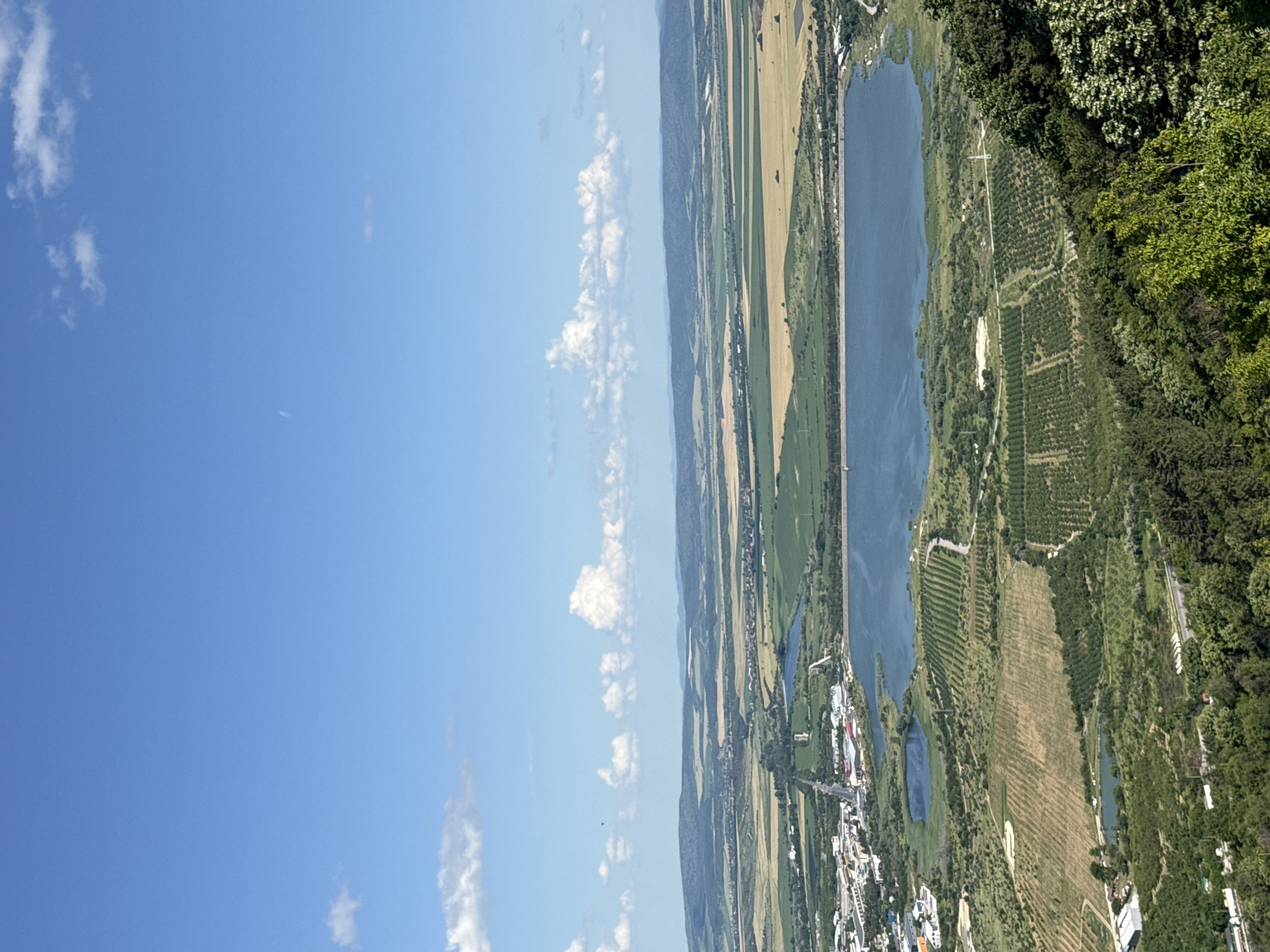 This is an aerial view photograph of a landscape near Shumen, Bulgaria. The image captures a vast expanse of land characterized by agricultural fields, a sizable body of water, and a distant, hazy mountain range. In the foreground, dense green trees and foliage dominate the lower portion of the frame. As the view recedes, the landscape transitions into neatly organized agricultural plots, some appearing as rows of crops and others as fields of varying colors, suggesting different types of vegetation or stages of growth. A prominent, dark blue lake or reservoir is centrally located in the mid-ground, with a dam or embankment visible along its edge. Adjacent to the water are some buildings and infrastructure, indicating a settled area or industrial zone. Further in the distance, rolling hills and mountains form the horizon, under a bright blue sky dotted with scattered white clouds. The overall impression is one of a rural or semi-rural setting, possibly a nature reserve or agricultural region on the outskirts of Shumen.