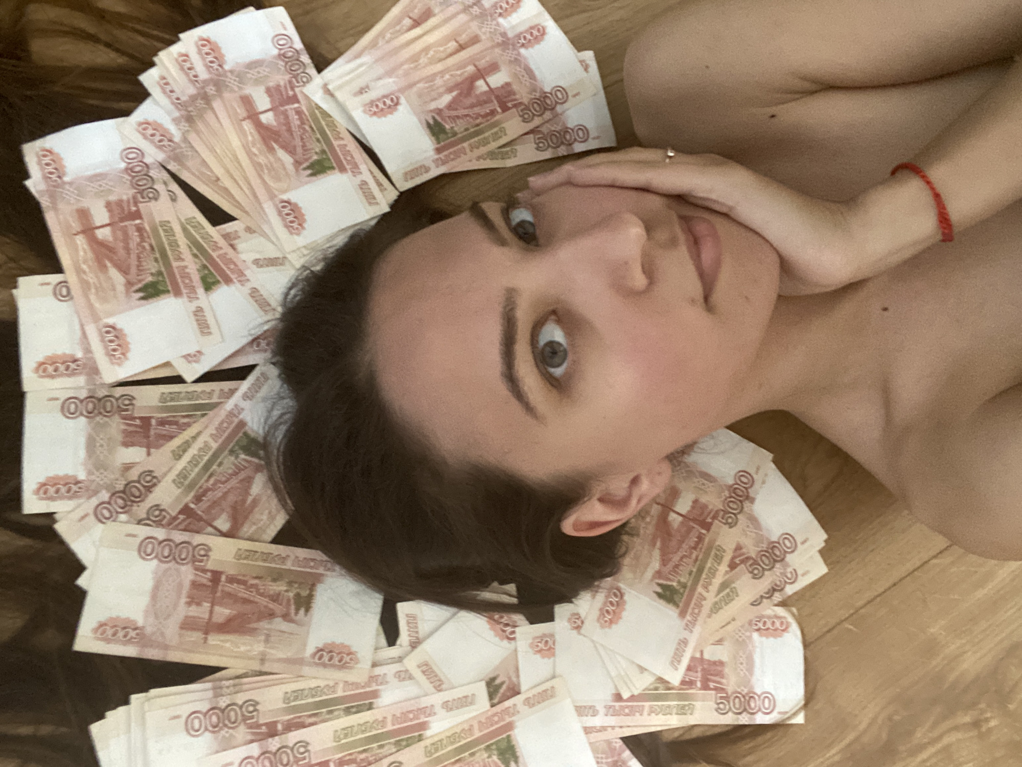 A woman is lying on the floor surrounded by Russian 5000 ruble banknotes. She is partially nude, with her head resting on some of the banknotes and her hand touching her face. The banknotes are scattered around her on what appears to be a wooden floor. The environment suggests an indoor setting, possibly a home. There are no details available to suggest this image's relevance to the city of Sevastopol, Ukraine.