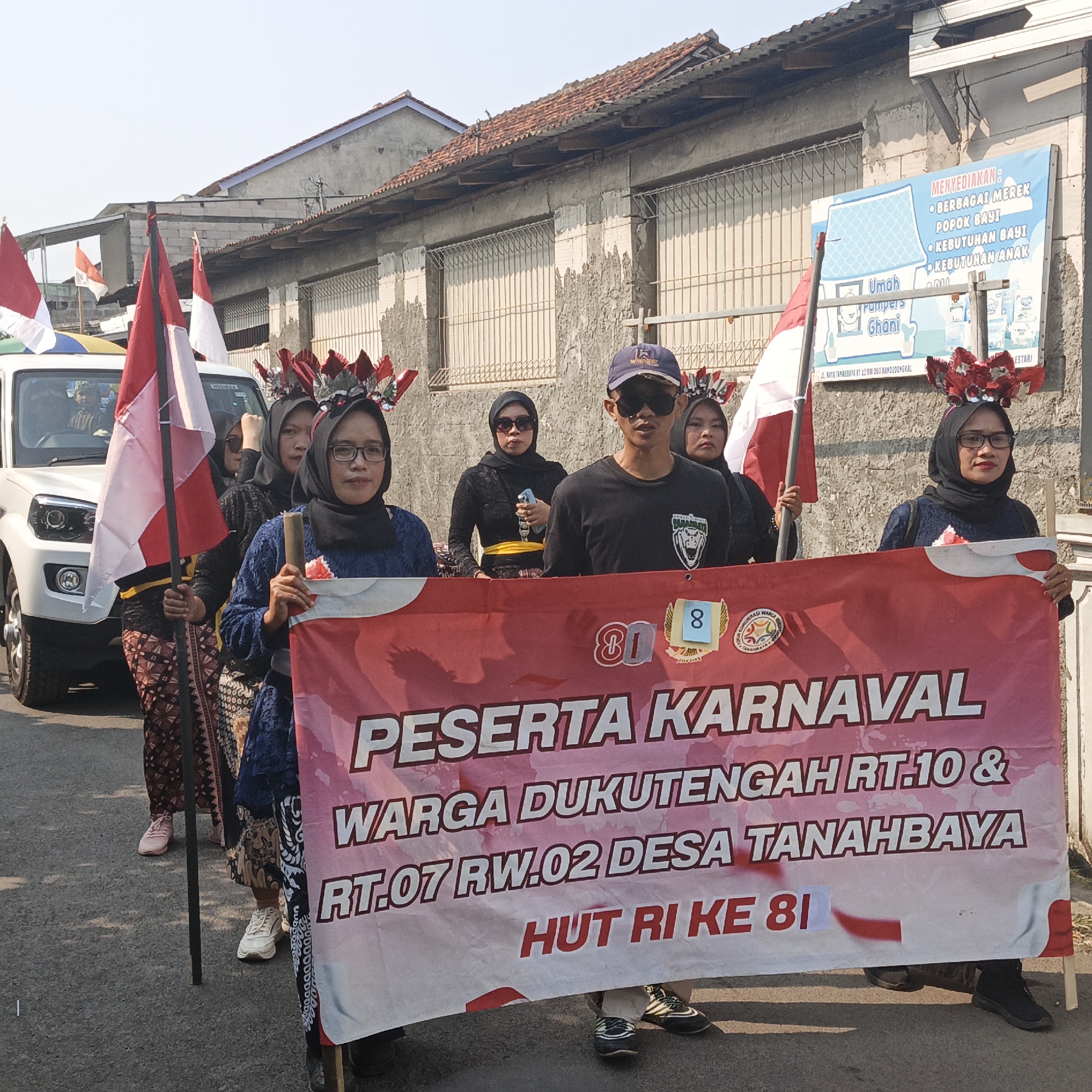 The image depicts an outdoor street scene, likely in Tanahbaya, Indonesia, featuring a parade or procession. In the foreground, a group of individuals, primarily women, are holding a large red and white banner with text in Indonesian. The banner reads "PESERTA KARNAVAL WARGA DUKUTENGAH RT.10 & RT.07 RW.02 DESA TANAHBAYA HUT RI KE 81", translating to "Carnival Participants of Dukutengah RT.10 & RT.07 RW.02 Tanahbaya Village 81st Indonesian Independence Day".
Several people are wearing traditional Indonesian attire, including batik skirts and headscarves. Some individuals are adorned with decorative headpieces. The people are walking on a paved street. Behind the banner carriers, a white SUV is visible, suggesting it might be part of the procession. Indonesian flags, red and white, are also present, held by some of the participants.
In the background, there are low-rise buildings with weathered facades and corrugated metal roofs. A light blue banner with text in Indonesian is attached to one of the buildings, advertising baby products. The sky is clear, indicating a sunny day. The overall impression is of a community event celebrating Indonesian Independence Day.