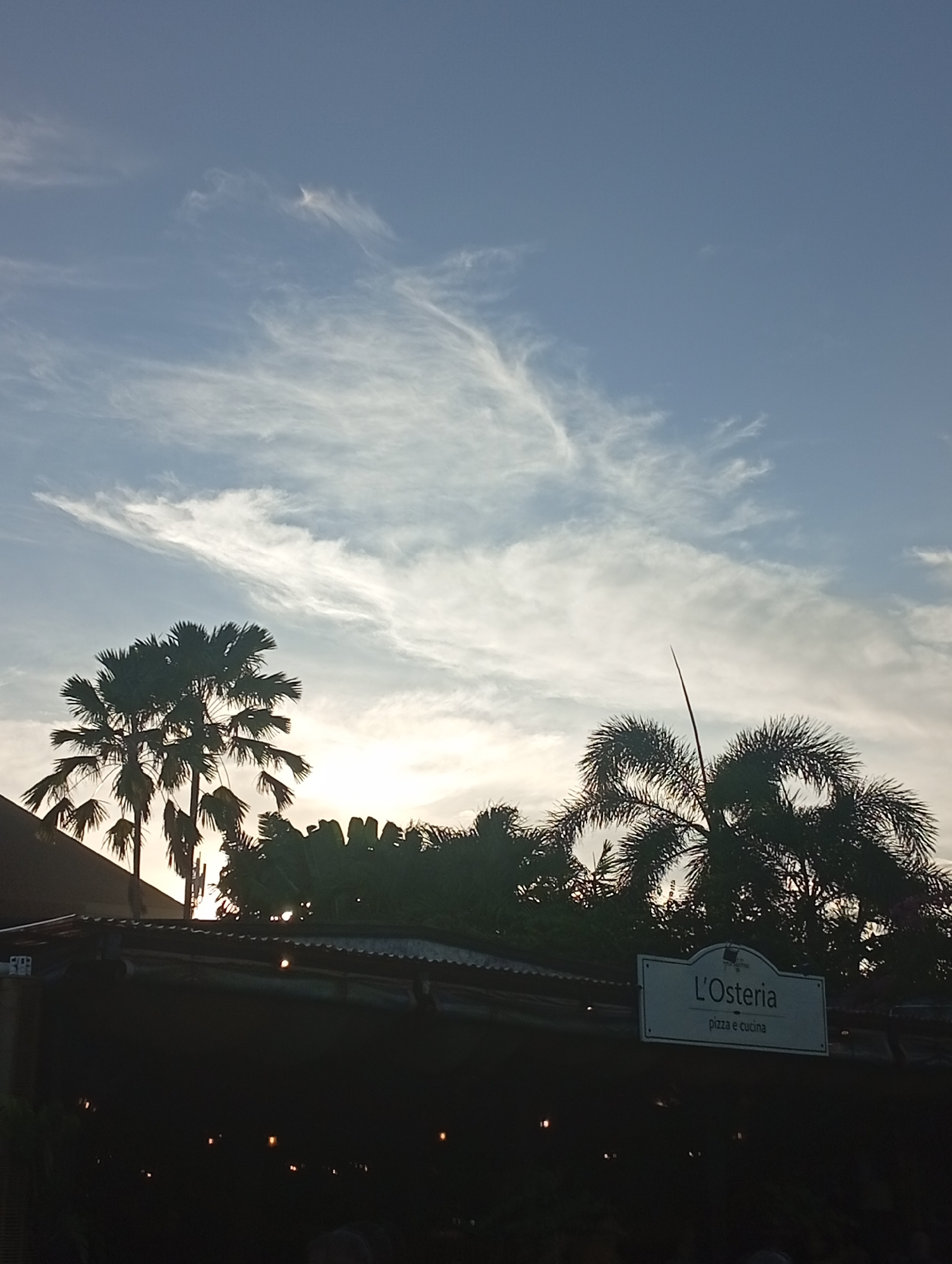 The image captures a serene tropical sunset over a restaurant, likely in Denpasar, Indonesia. The sky dominates the upper portion, displaying a soft blue hue adorned with delicate, wispy white clouds that stretch across the frame. A warm, golden glow from the setting sun peeks over the horizon, largely obscured by the silhouettes of lush palm trees. On the left, tall palms with broad, fan-shaped fronds stand prominently, while on the right, trees with feathery leaves are visible. Below the tree line, the dark outline of a building or outdoor dining area emerges. A clear white sign is mounted on this structure, bearing the Italian text "L'Osteria" above "pizza e cucina". Faint, warm-toned lights glow indistinctly in the dark foreground, hinting at an evening ambiance. No discernible people or specific activities are visible. The overall scene conveys a tranquil, late afternoon or early evening mood.