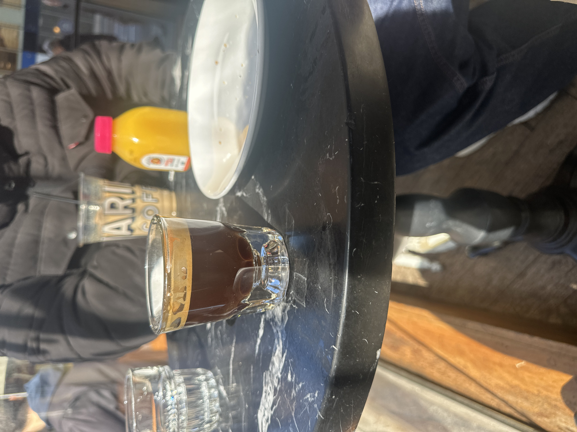 An eye-level shot shows a small glass of dark liquid (possibly coffee or espresso), a small glass, a bottle of juice, and a plate on a round, dark marble-patterned table. A person in a dark jacket is sitting at the table. The table is likely located outdoors, with a hint of a wooden floor in the background. The location is in an outdoor cafe/restaurant with the time during the daylight. The location is not discernible as a specific location in the city of New York, United States of America.