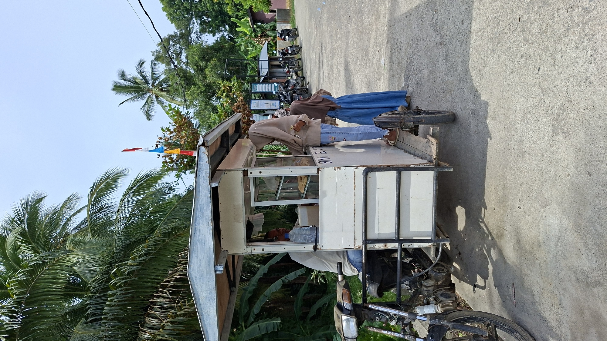 A mobile food cart, resembling a small, white, rectangular booth mounted on a motorcycle-drawn trailer, is parked on a paved road in Asan, Indonesia. The cart has a silver corrugated metal roof and a display window with a glass front. Inside the cart, a man in a light-colored shirt and blue jeans is seated, with a plastic bottle of water on the counter. Two young women are interacting with the vendor. One woman, wearing a light brown hijab and blue jeans, stands to the right of the cart and appears to be ordering or receiving food. The other woman, wearing a dark brown hijab and a blue dress, stands beside her. In the background, several parked motorcycles are visible, along with lush green tropical vegetation, including palm trees, and a few buildings. Small, colorful flags are visible on a pole in the upper left portion of the image. The scene depicts a typical street food vendor operation in a tropical setting.