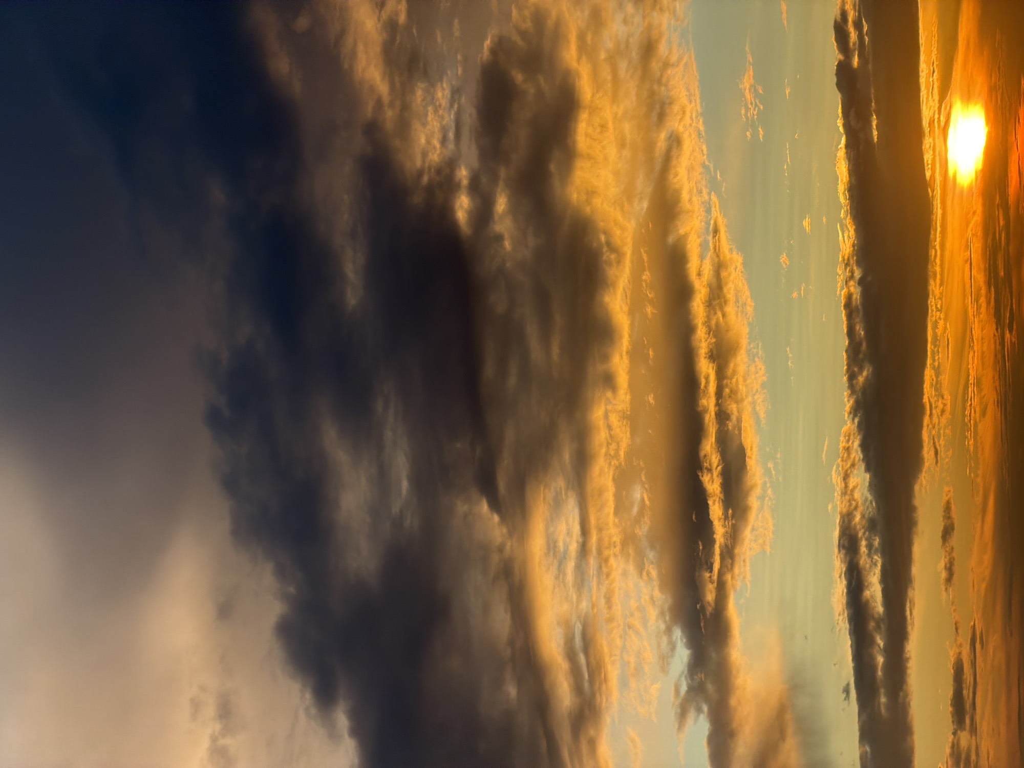This is a vertical, full-frame photograph of a sunset over an unknown location. The sky is filled with dramatic clouds, ranging in color from deep charcoal and dark gray to bright golden orange and yellow. The sun, a brilliant white orb, is visible near the bottom right corner of the frame, casting a warm glow. The clouds are textured and layered, with some appearing wispy and others dense and heavy. The overall impression is one of a powerful and vibrant natural display. There are no people, structures, or any identifiable man-made objects in the image.
