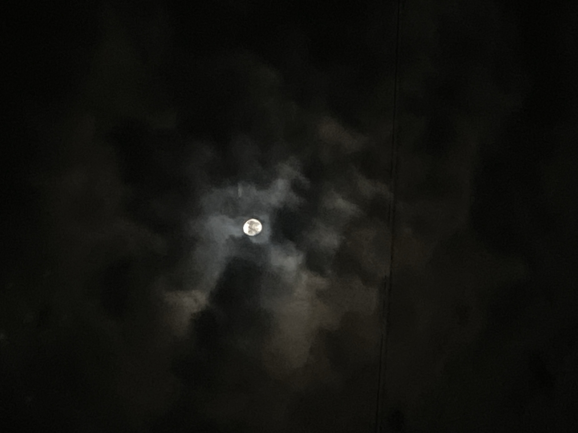 The image captures a night scene featuring a prominent full or near-full moon, partially obscured by dark, swirling clouds. The moon, a bright circular disk, is centrally located and glows intensely, illuminating the edges and translucent sections of the surrounding cloud cover. These clouds vary in density, with lighter, more ethereal formations directly around the lunar surface and darker, more opaque masses filling the majority of the night sky. The overall impression is one of deep darkness, characteristic of nighttime. A subtle, dark, vertical line, possibly a wire or a distant structural element, is faintly visible on the right side of the frame, extending downwards into the darkness. No specific activity or event is depicted beyond the natural celestial display. The scene conveys a serene yet atmospheric mood, set against the backdrop of the night sky in Chaikovsky, Russia. There is no visible text in the image.