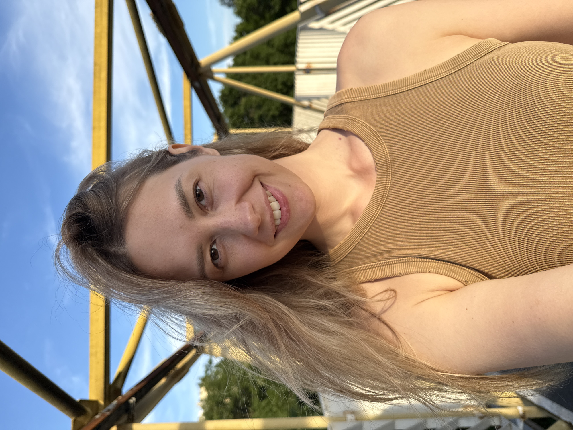 A young adult Caucasian female is depicted in an outdoor setting, likely a selfie. She has light blonde or light brown, somewhat wind-swept hair, and a fair complexion with visible pores, freckles, and a small mole on her upper chest and right arm. Her dark brown eyes are directed at the viewer, and she exhibits a broad smile, revealing her upper teeth. She is wearing a fitted, ribbed tank top in a tan or light brown color.
The background features prominent yellow-painted metal truss structures, indicative of a bridge or an overhead walkway, with some visible weathering and darker patches. A clear blue sky with minimal white clouds is visible above the structures. Green foliage from trees is present in the middle ground, and a white corrugated fence or wall runs horizontally in the distant background. The lighting suggests natural sunlight, illuminating her right side. This scene is located in the city of Nebug, Russia.
