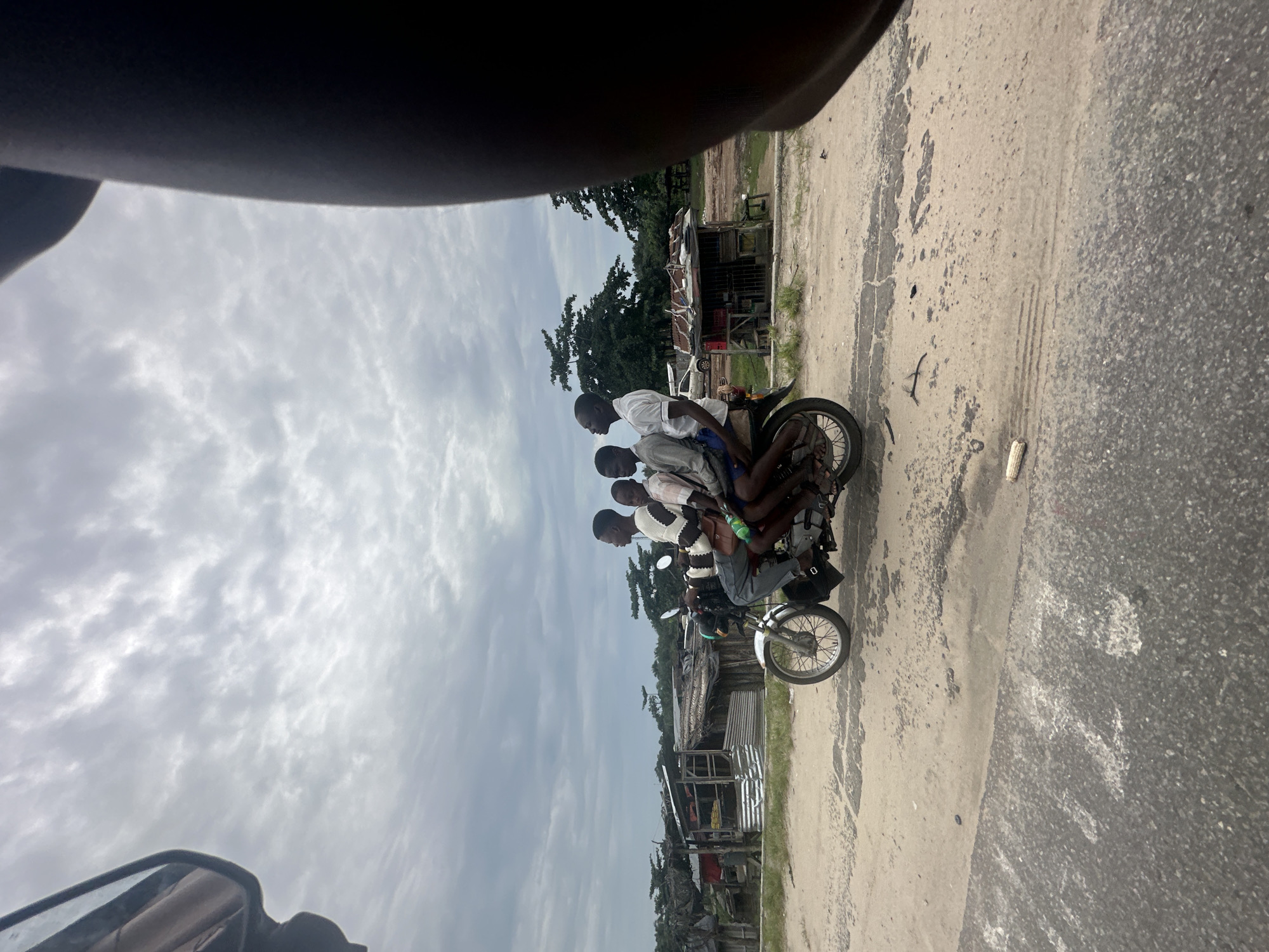 Four young men are riding on a single motorcycle on a dusty road in Badagry, Nigeria. The rider in the front, wearing a white and brown striped sweater, is looking forward. Behind him, three passengers are seated, also looking forward. The motorcycle is a silver and red model. The background features a village with small wooden structures, some with corrugated metal roofs, and lush green trees. The sky is overcast with clouds. The scene appears to be a typical mode of transportation in the area.