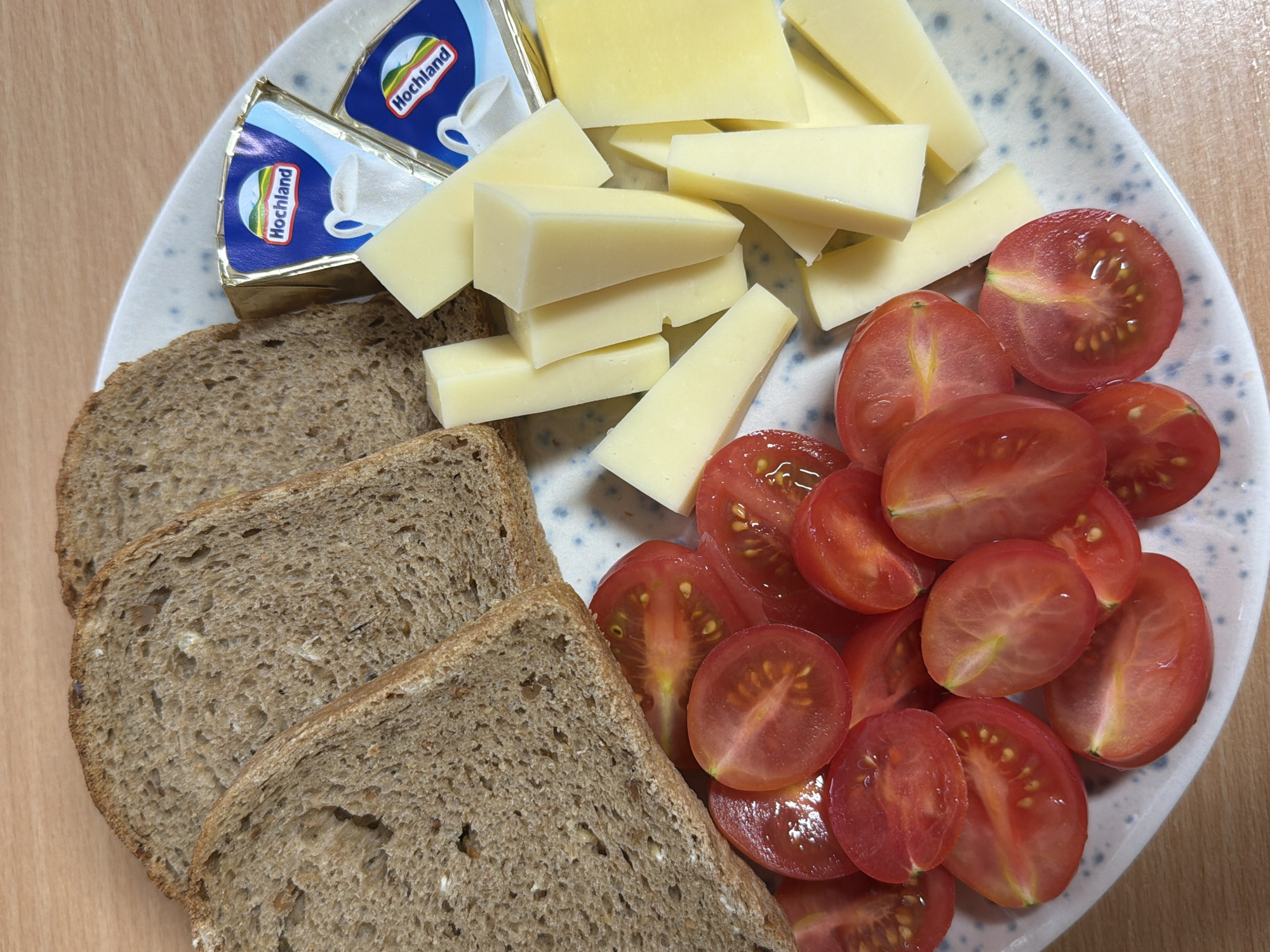 A top-down shot captures a simple meal or snack arranged on a white plate with a subtle blue speckled design, resting on a light brown wooden surface. The plate is divided into sections by the various food items.
In the lower left, three slices of dark, coarse-grained bread, likely whole grain or rye, are neatly stacked. The bread appears robust with visible seeds or grains.
Towards the top left, two individual portions of spreadable cheese are presented in their blue and gold foil packaging. The brand name "Hochland" is clearly visible in white text on the blue background, accompanied by a logo featuring a mountain and a white cup.
Adjacent to the bread and cheese packaging, a generous amount of yellow cheese is arranged. The cheese is cut into various shapes, predominantly triangular wedges and rectangular sticks, suggesting a firm, semi-hard variety.
Filling the right side of the plate are numerous halved red cherry tomatoes. Their vibrant red color and juicy interiors with visible seeds contrast with the lighter tones of the other foods. The cut surfaces of the tomatoes show their watery texture.
The overall scene depicts a fresh and simple food platter, possibly prepared for breakfast, lunch, or a light snack. While the provided context suggests the location is Sofia, Bulgaria, there are no specific visual cues within the image itself to confirm this, other than the German brand Hochland, which is widely available internationally. The time of day and weather are not discernible.