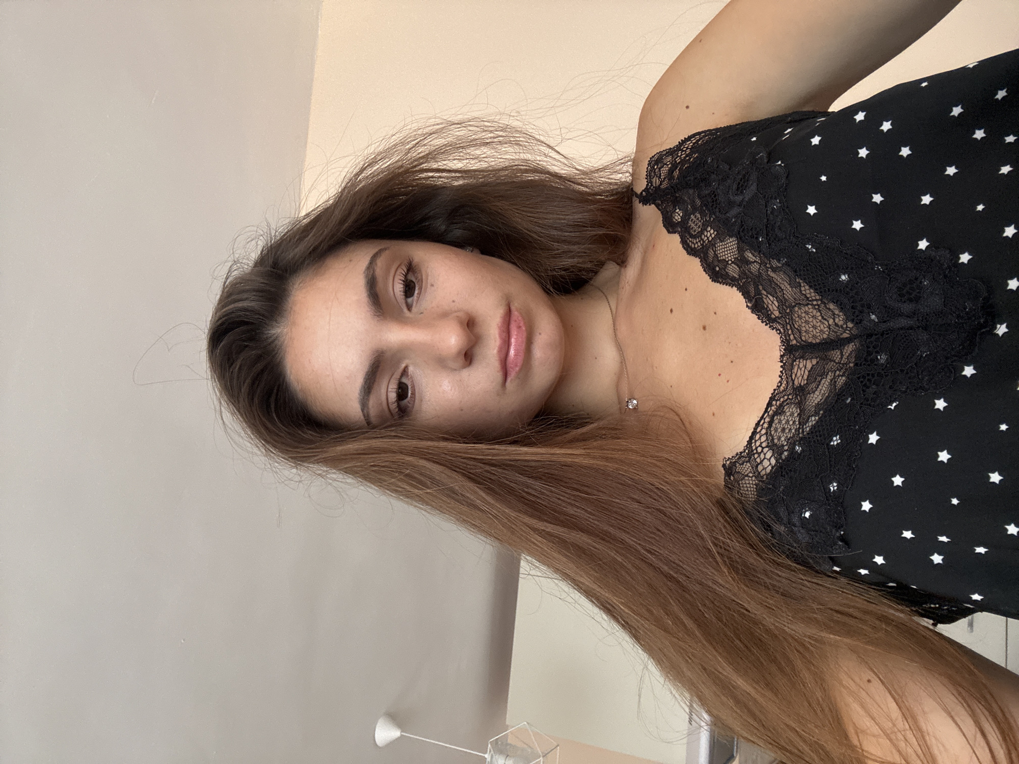 A young woman with long, light brown hair and brown eyes is looking down at the camera. She is wearing a black camisole with white stars and black lace trim. She has a necklace with a small pendant. The background is a plain, light-colored wall. The woman is positioned in what appears to be an indoor setting. There are no discernible landmarks or features that would pinpoint a specific location within the city of Varna, Bulgaria, based solely on the visual information provided in the image.