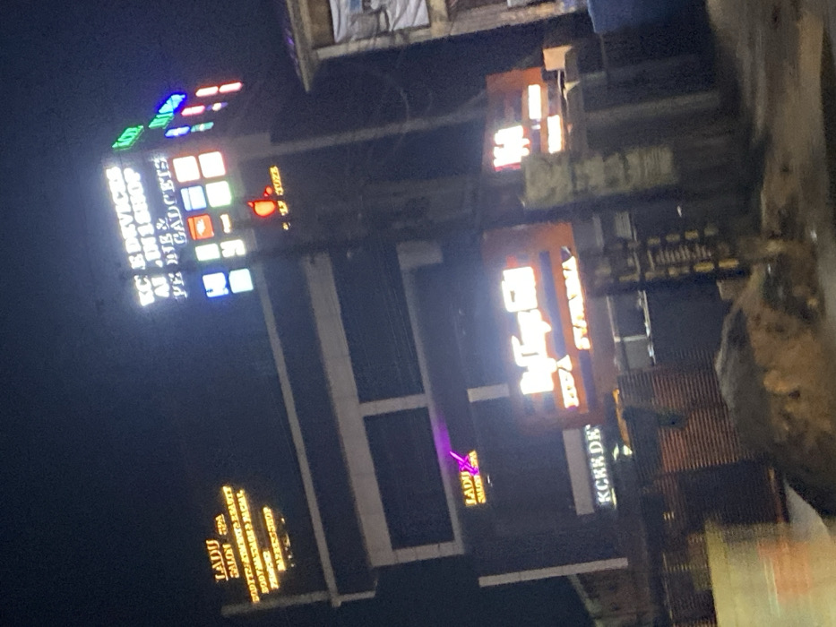 Nighttime view of a tall building adorned with illuminated signs. “KC E Devices Al In 1 Shop Ye Me & Gadgets” is stacked vertically. Below, signs for “Ladu Salon Spa” and “KCKE DE” can be discerned. An adjacent sign features a stylized text “HEDA” and what appears to be "SWAXHA." The lighting casts a vibrant glow, adding to the dynamic energy of the cityscape.