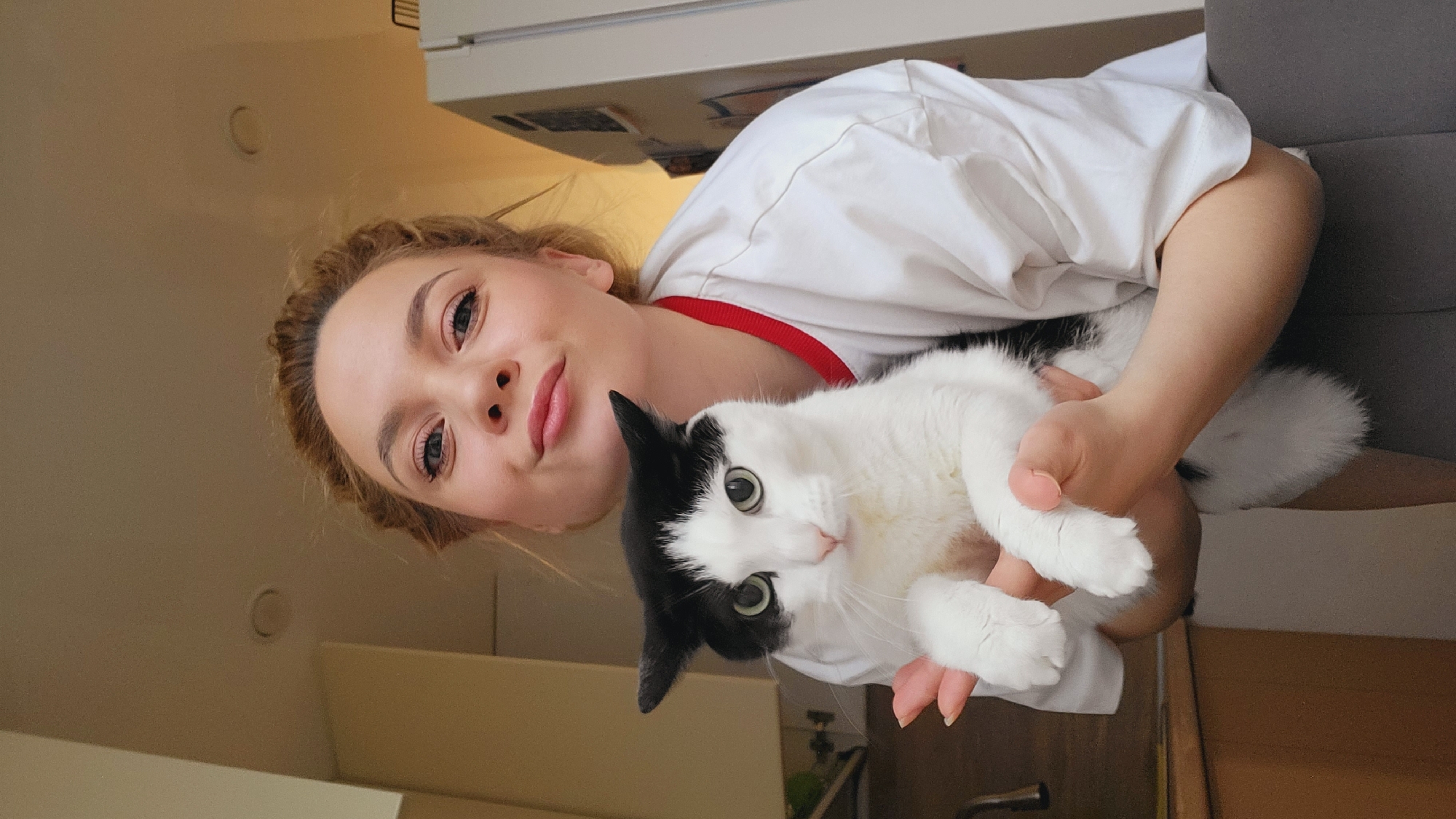 A woman holds a black and white cat in an indoor environment. The woman has fair skin, blonde hair, and is wearing a white shirt with red trim around the collar. She is looking toward the camera. The cat has green eyes and a white body with black markings on its head. The background is a mix of household elements, including a white refrigerator and hints of a light-colored wall. The location is Chaikovsky, Russia, based on the provided information.